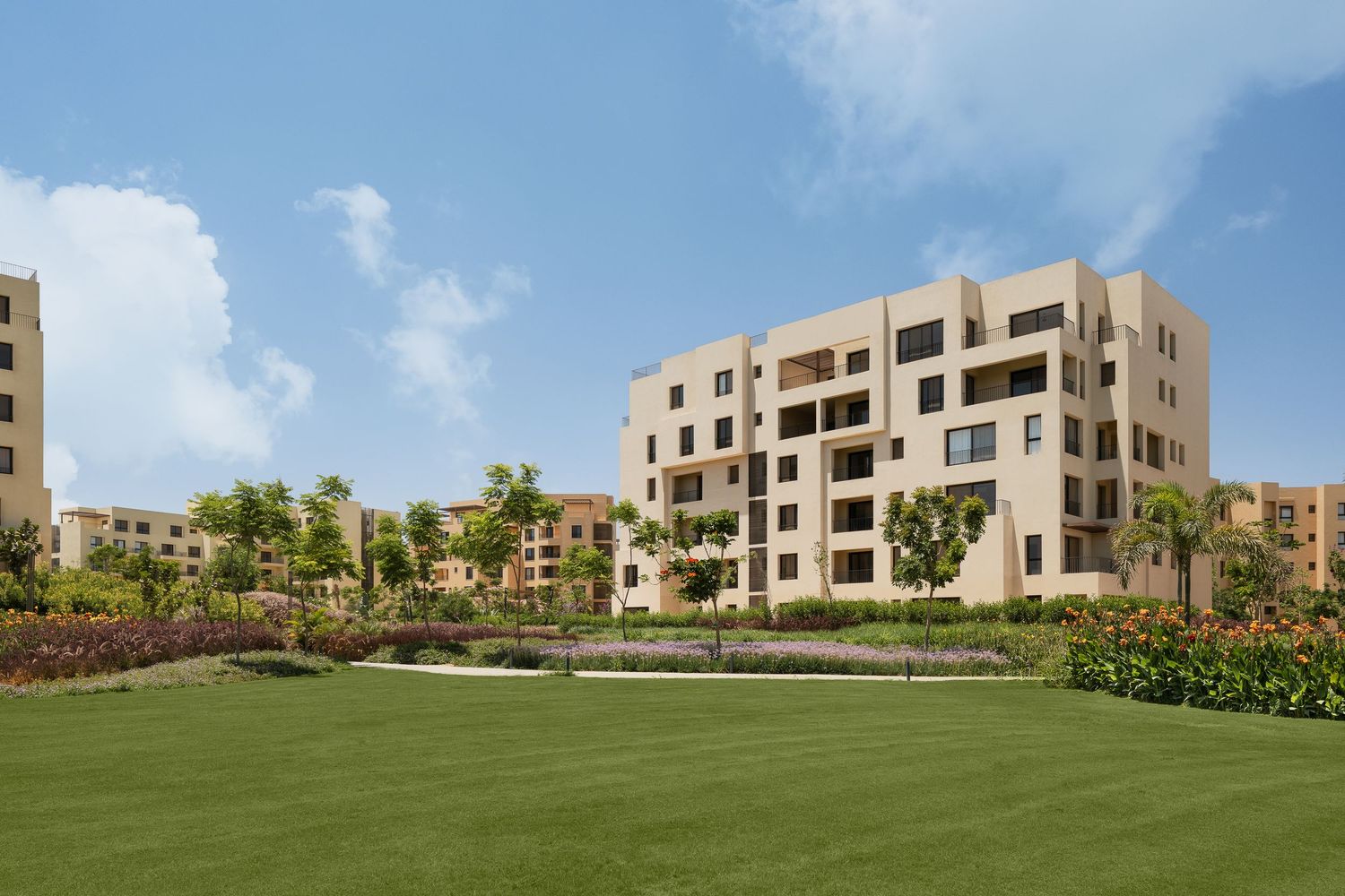 Luxury residential apartment complex with beige facades surrounded by lush green lawns and tropical landscaping under blue sky.