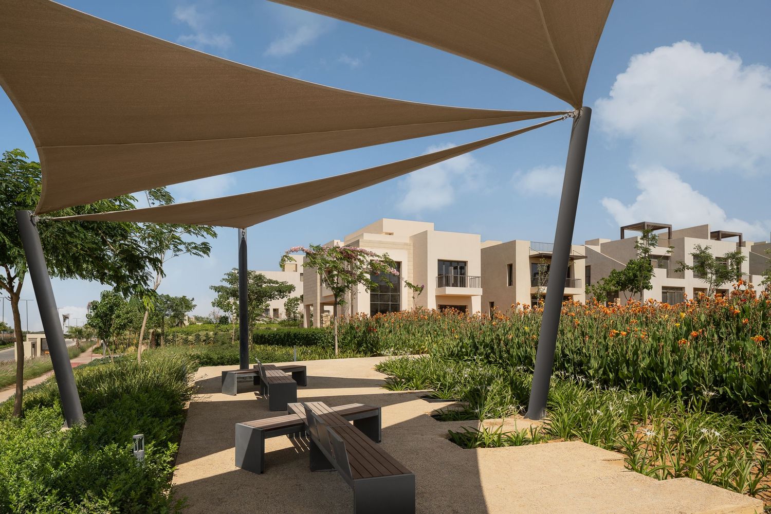 Shaded outdoor lounge area with sail canopies, stone benches, and vibrant gardens beside modern townhouses.