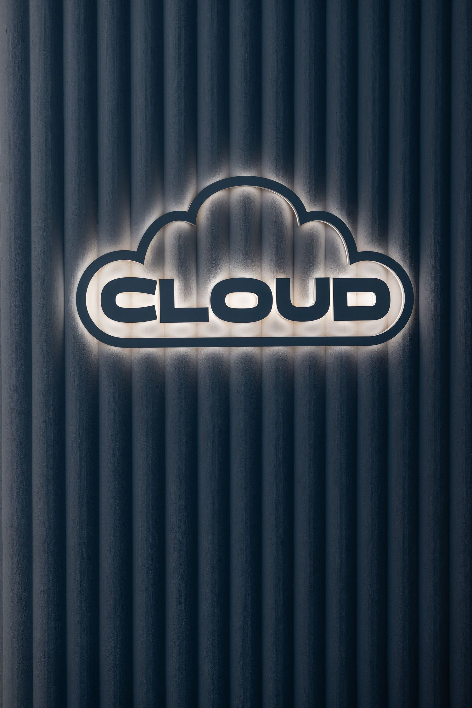 Illuminated cloud-shaped logo sign with halo backlight effect mounted on dark ribbed metal wall panel.