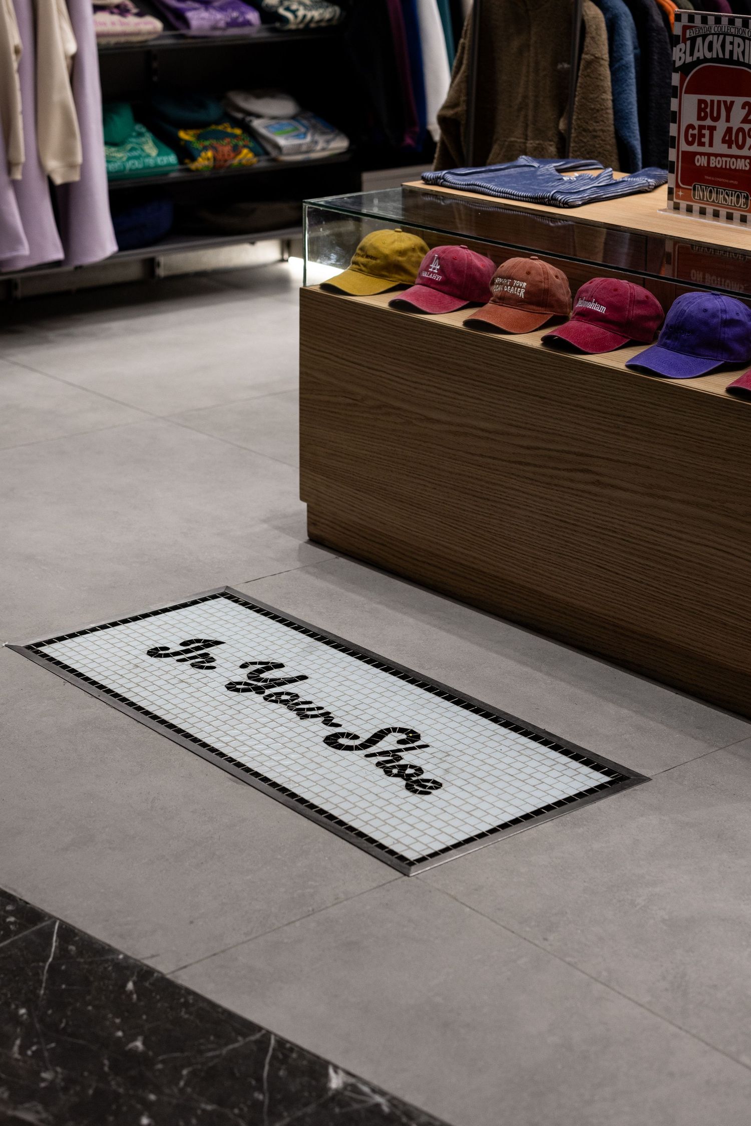 Retail store floor featuring mosaic tile mat reading In Your Shoe with colorful caps and apparel displayed on wooden fixture.