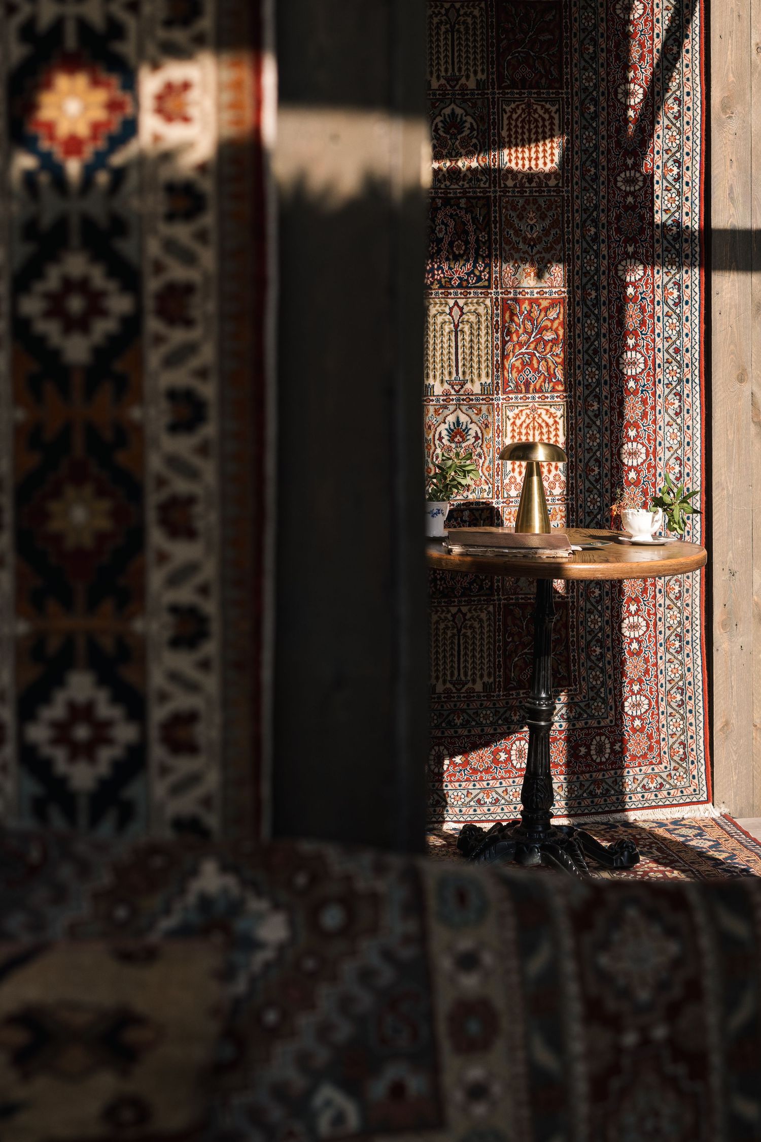 Sunlit room framed by Persian rugs revealing small table with brass lamp through patterned textile doorway.