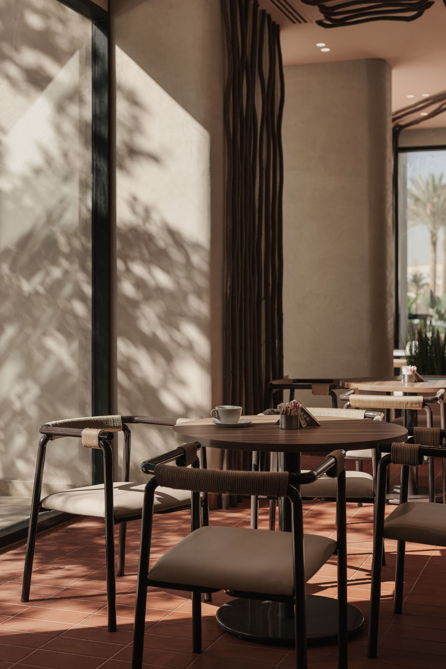 Sun-drenched cafe dining area with black metal chairs, round tables, terracotta floors, and leaf shadow patterns.