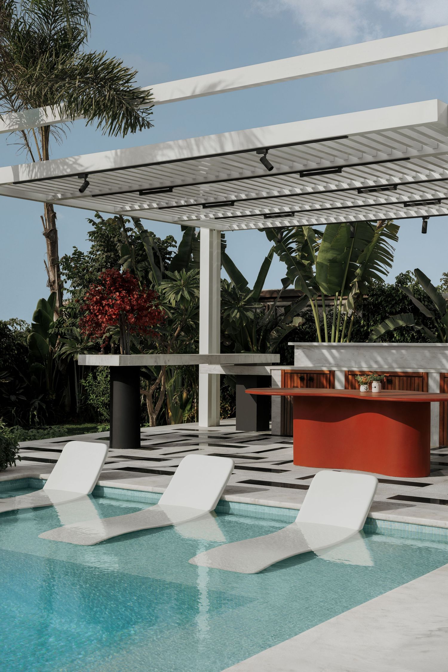 Modern poolside pergola with white sun loungers in water, red curved bar, and lush tropical landscaping.