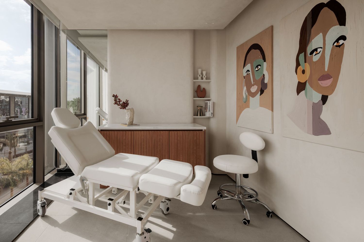 Bright spa treatment room with a white massage table, modern artwork, and floor-to-ceiling windows with city views.