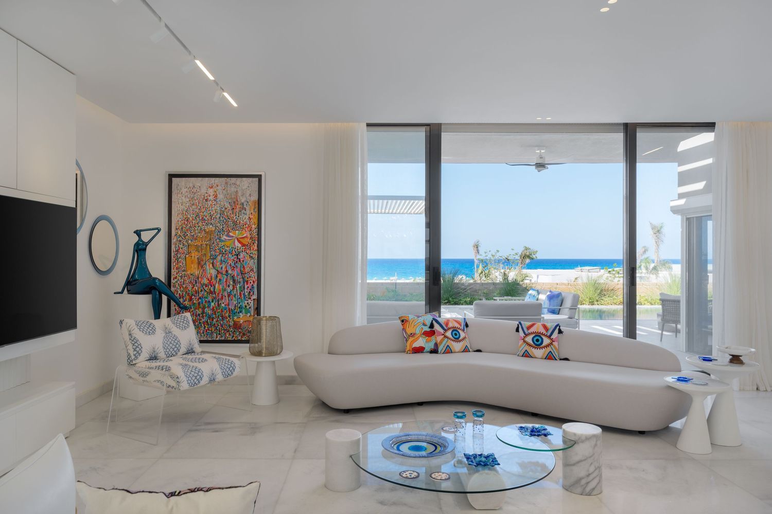 Bright white living room with curved sofa, vibrant artwork, blue glass coffee table, and panoramic ocean view.