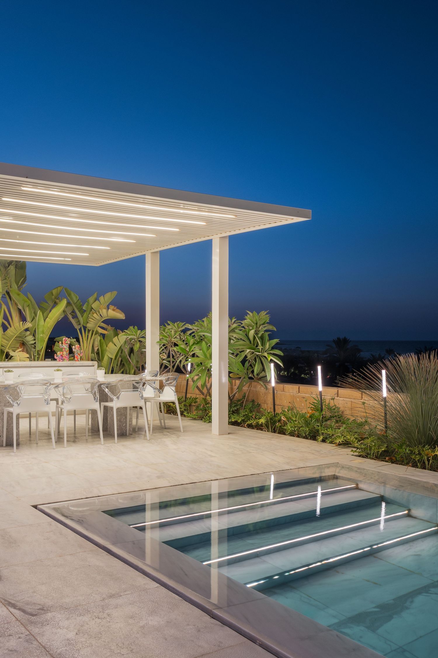 Illuminated outdoor pergola with dining area beside a stepped pool at twilight with ocean backdrop.
