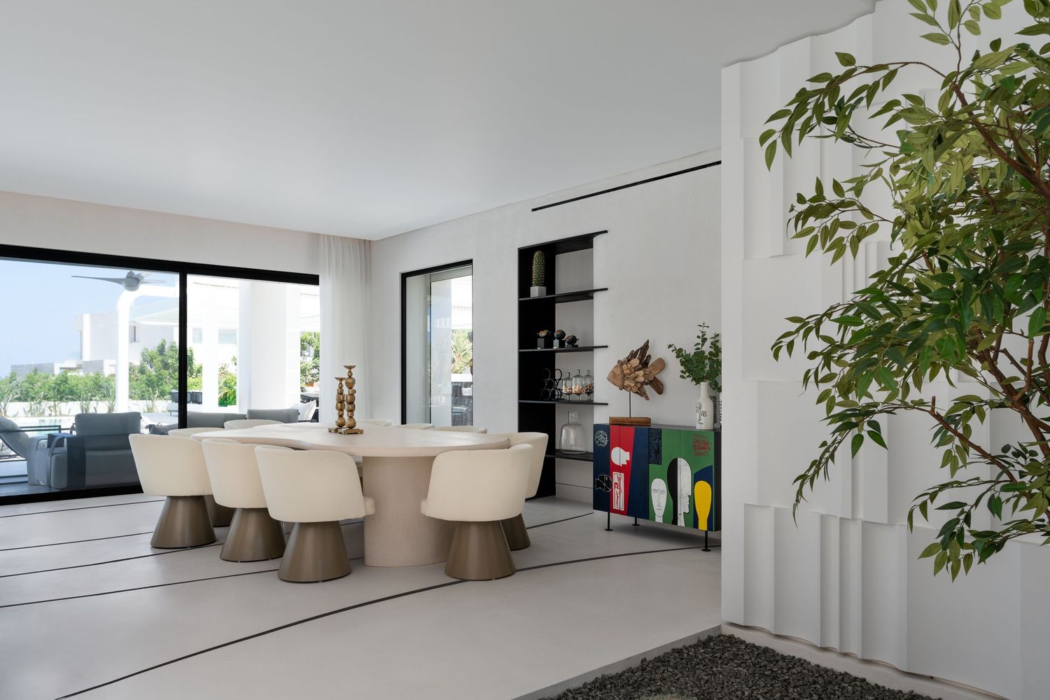 Bright modern dining area with cream chairs, wooden table, indoor tree, and colorful bookshelf display.