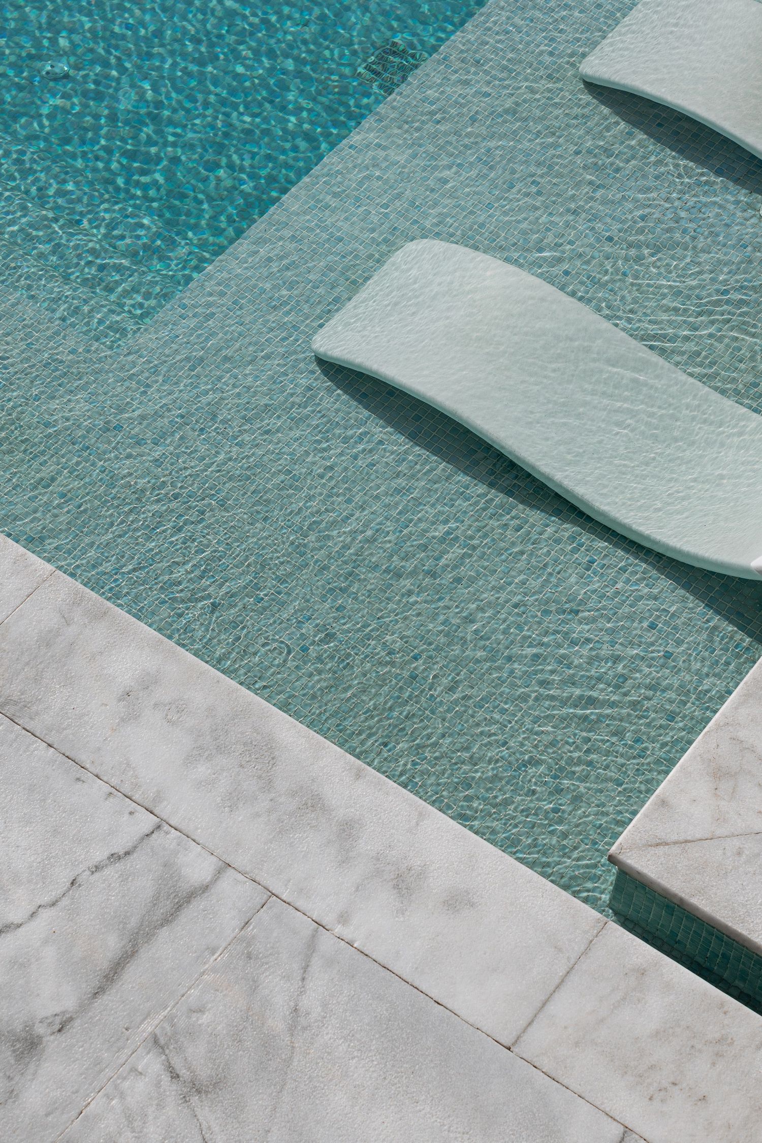 Aerial view of a sunken lounge chair in a turquoise pool with mosaic tiles and white marble pool surround.