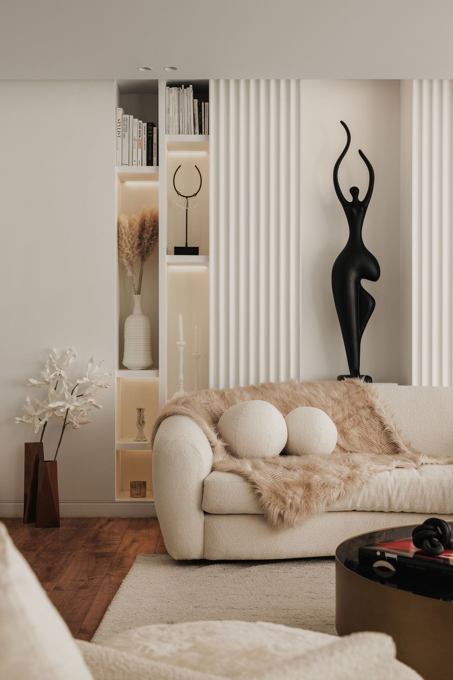 Modern living room featuring cream bouclé sofa, fur throw, black female figurine sculpture, and illuminated shelving.