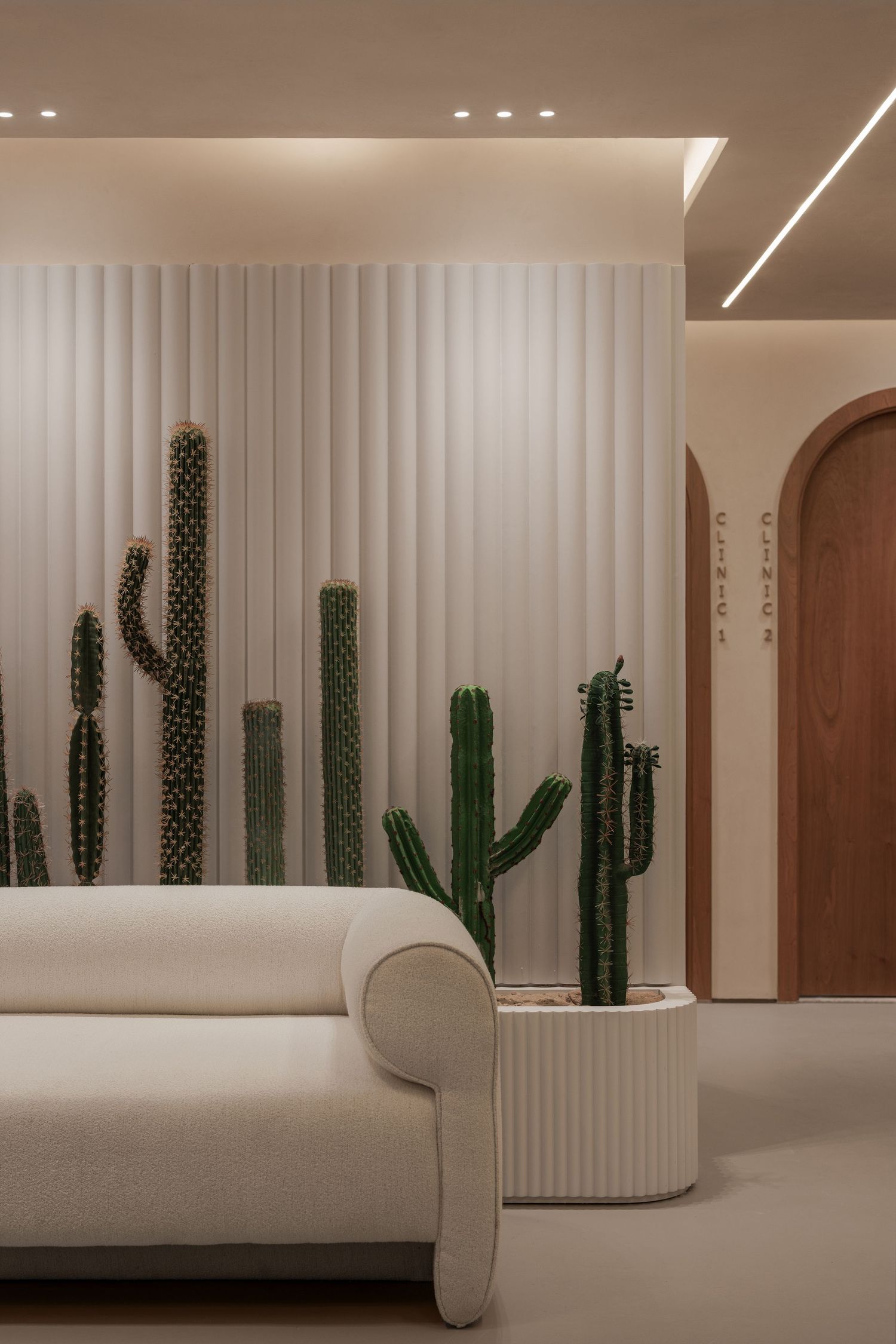 Modern spa lobby featuring tall cacti against white fluted wall panels, cream curved sofa, and warm recessed lighting.