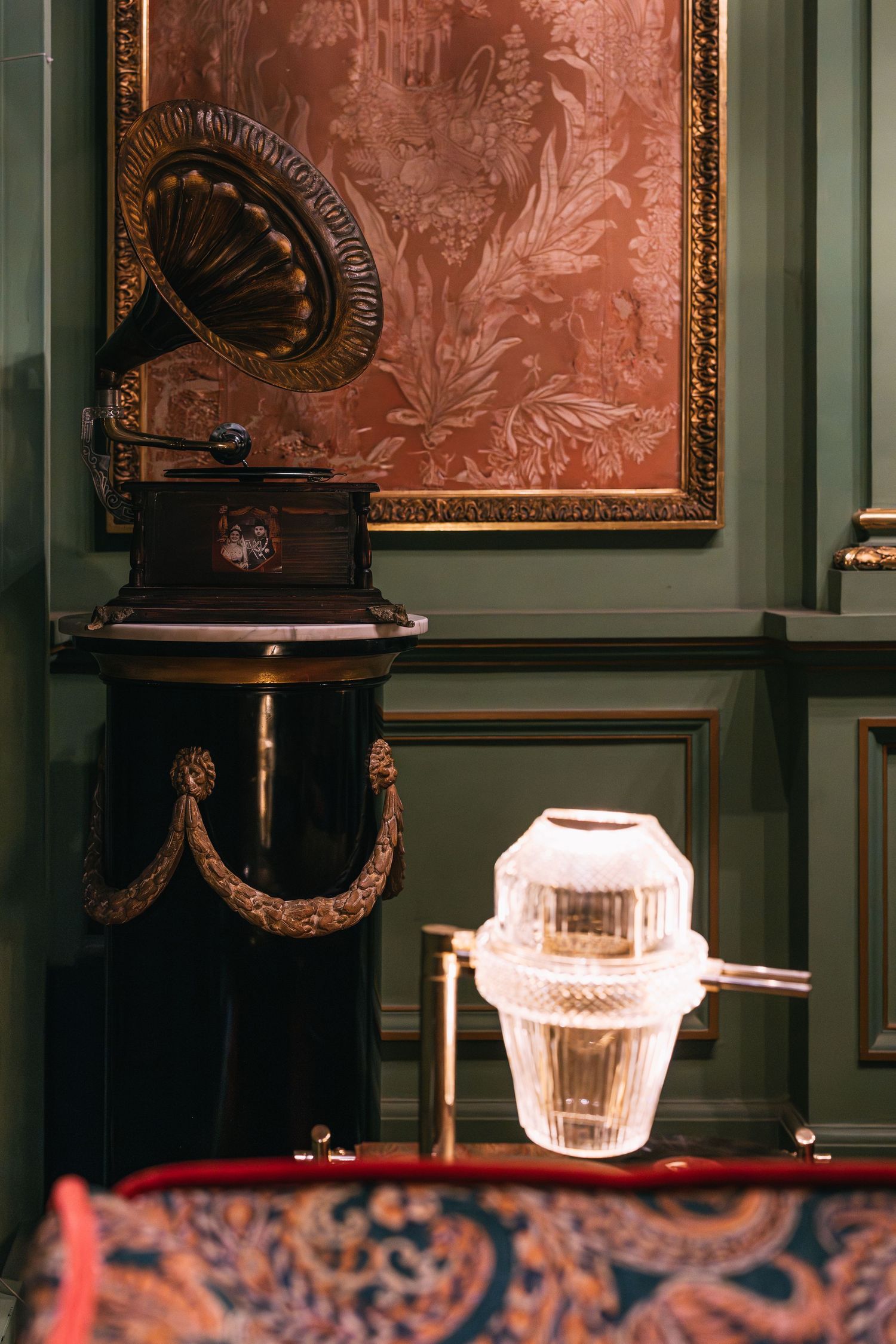 Antique gramophone beside a crystal glass lamp on an ornate black pedestal against green paneled wall.