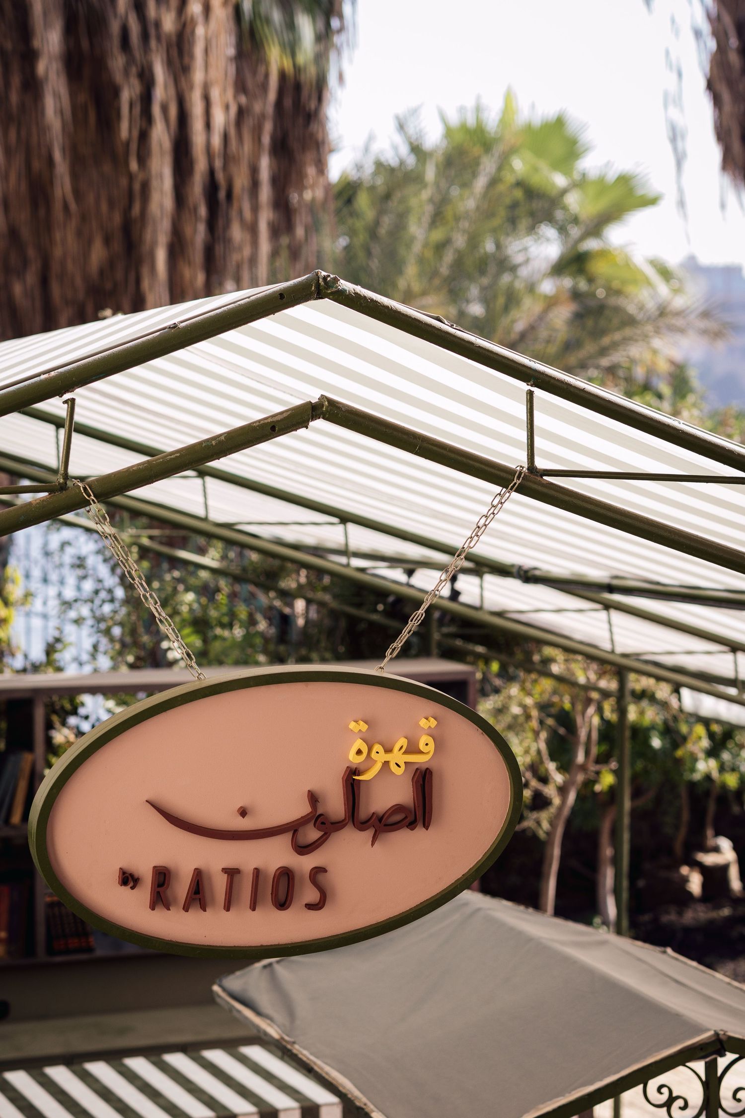Oval hanging sign for Ratios cafe with Arabic script displayed under striped awning surrounded by palm trees.