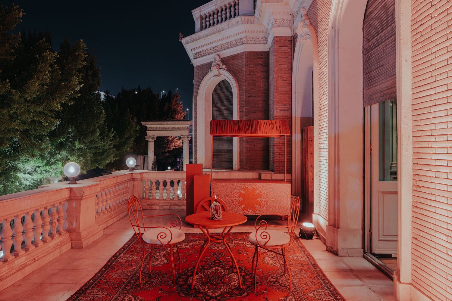 Night terrace with ornate iron furniture, red Persian rug, and brick building facade under warm lighting.
