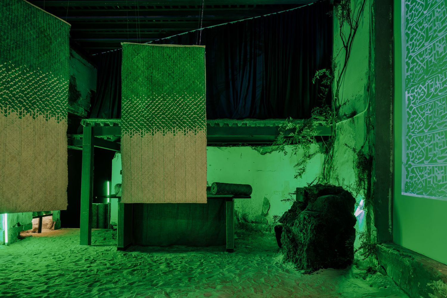 Eerie green-lit installation art space with hanging textile panels and rocky debris on sandy floor.