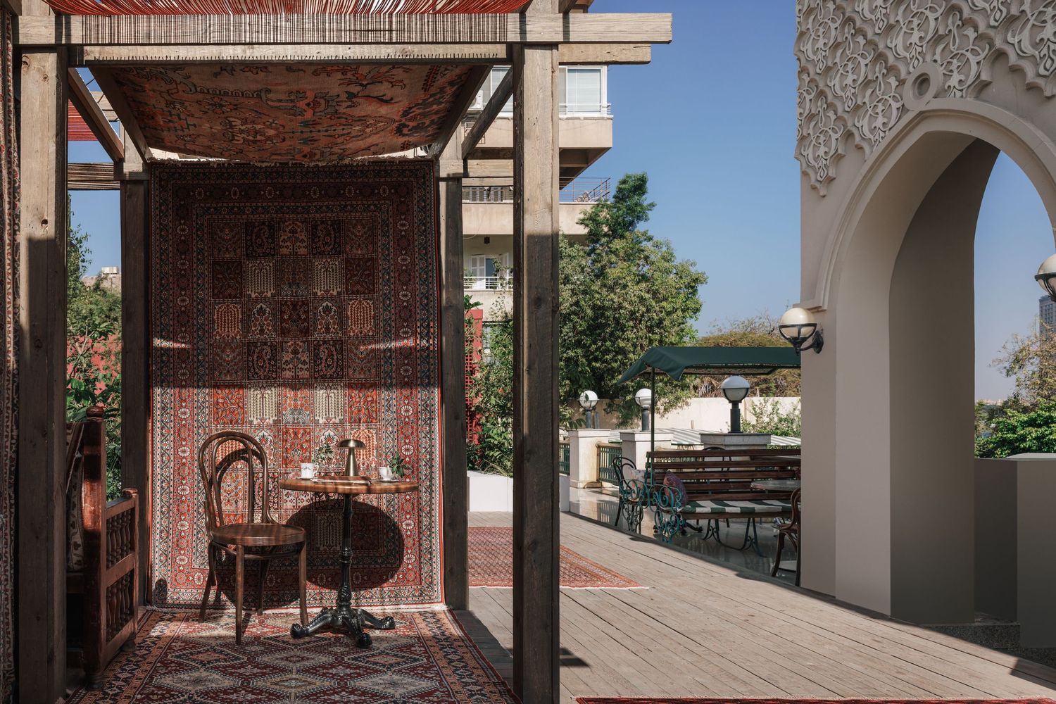 Ornate Persian rugs covering a wooden outdoor pavilion with chairs beside a decorative arch.