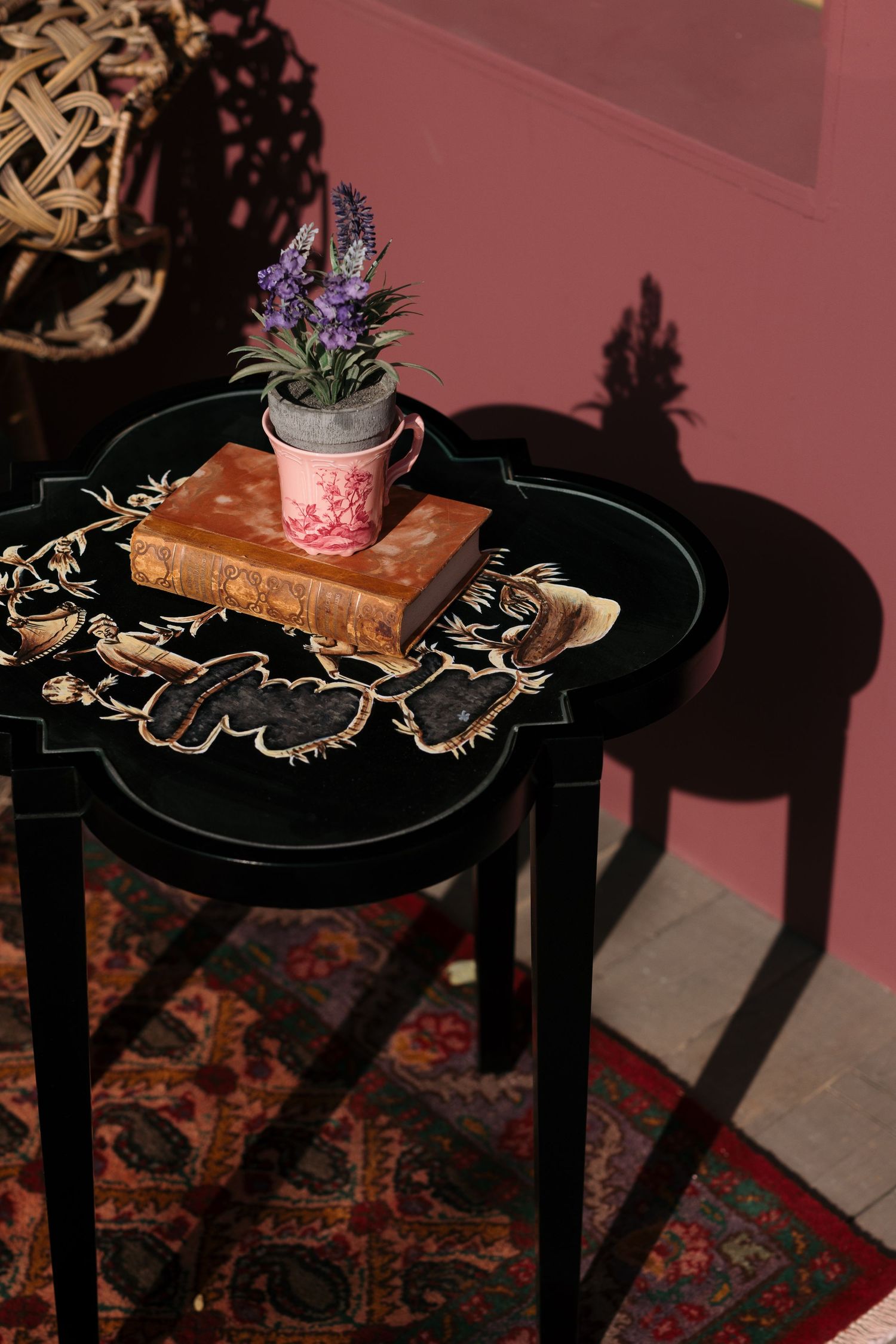 Black side table with ornate tray holding an antique book, lavender plant in pink pot, casting shadows on pink wall.