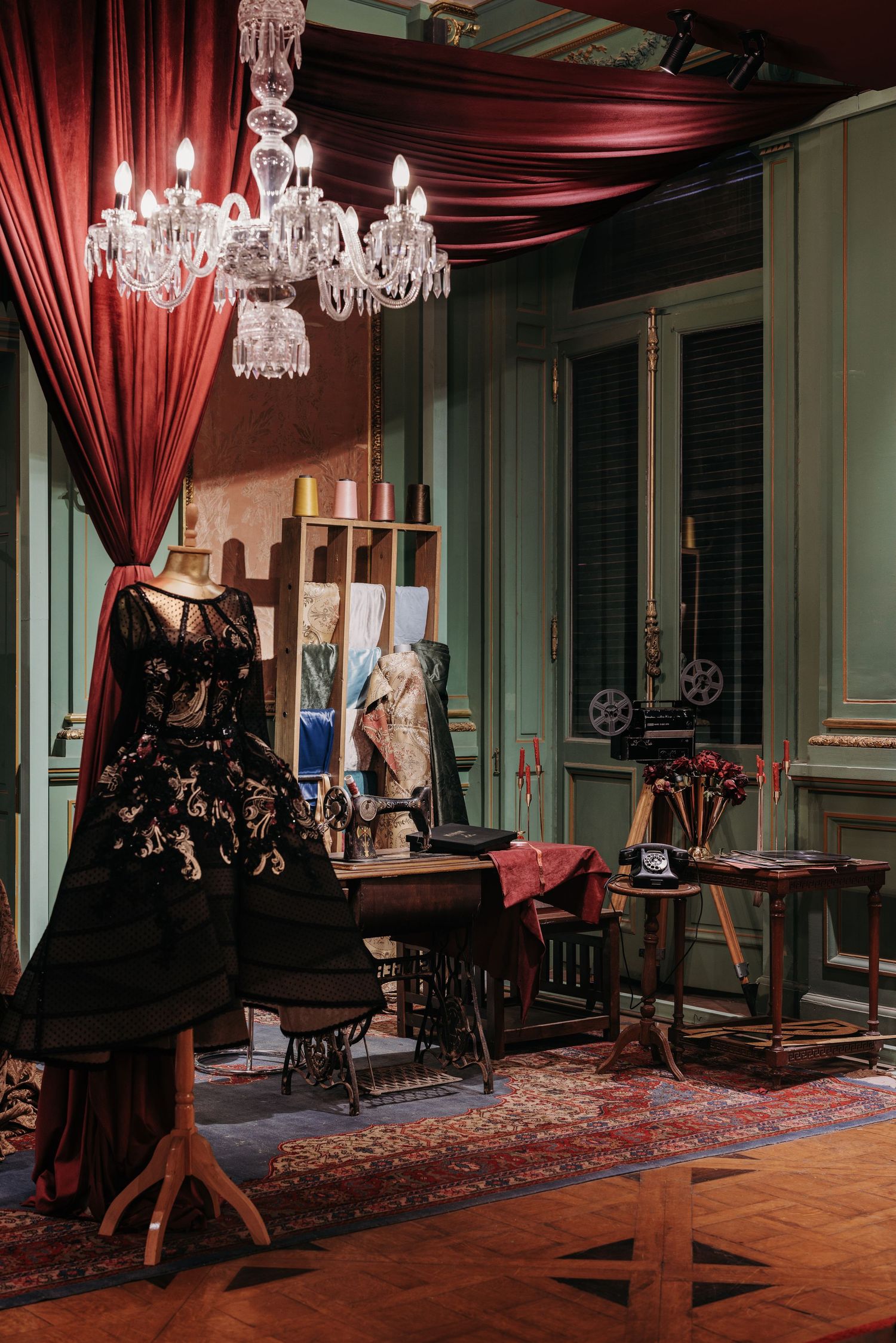 Elegant vintage atelier with black floral gown on mannequin, crystal chandelier, and red drapes.