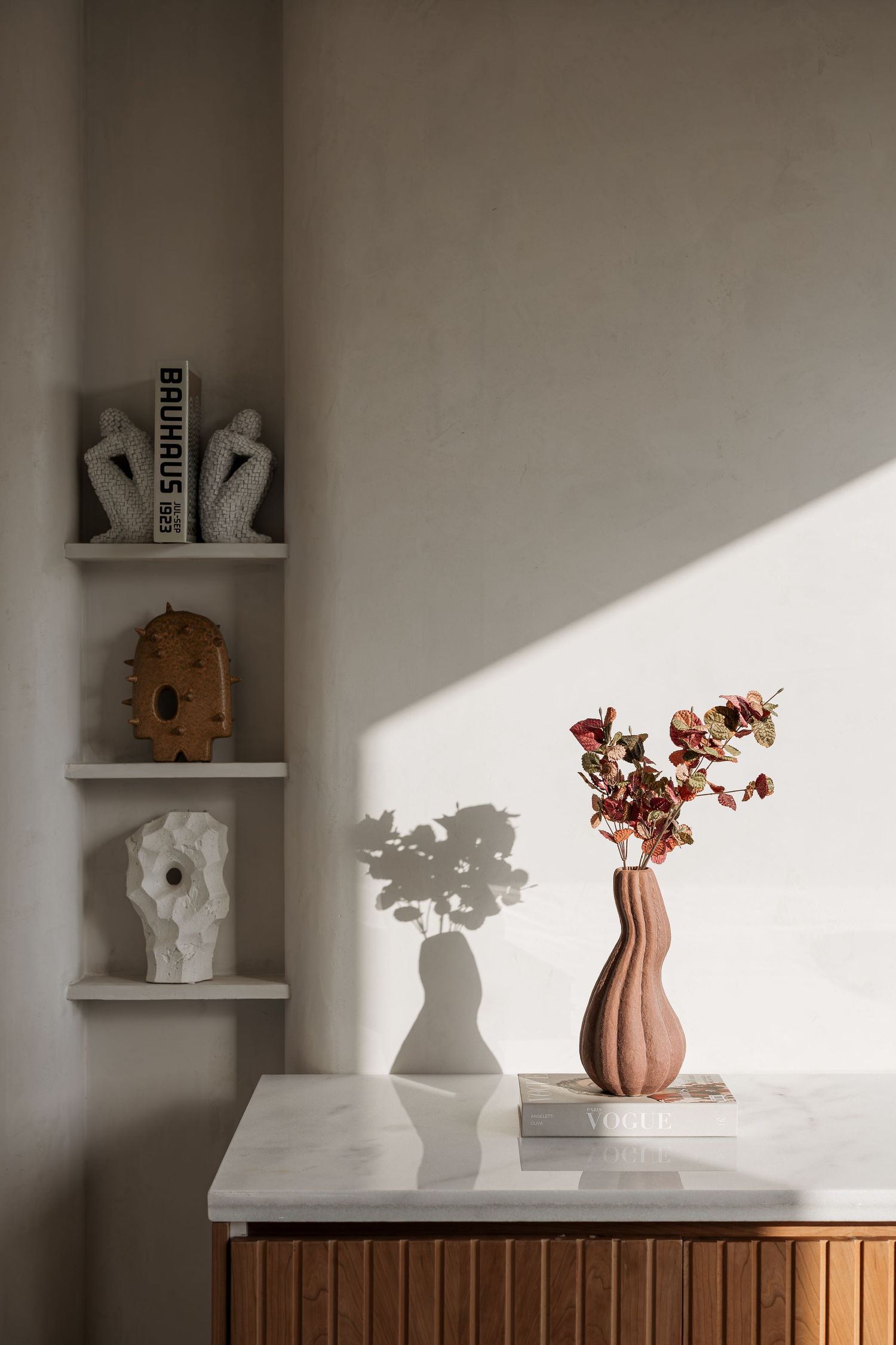 Terracotta wavy vase with dried flowers on marble countertop beside sculptural decor on minimalist wall shelves.