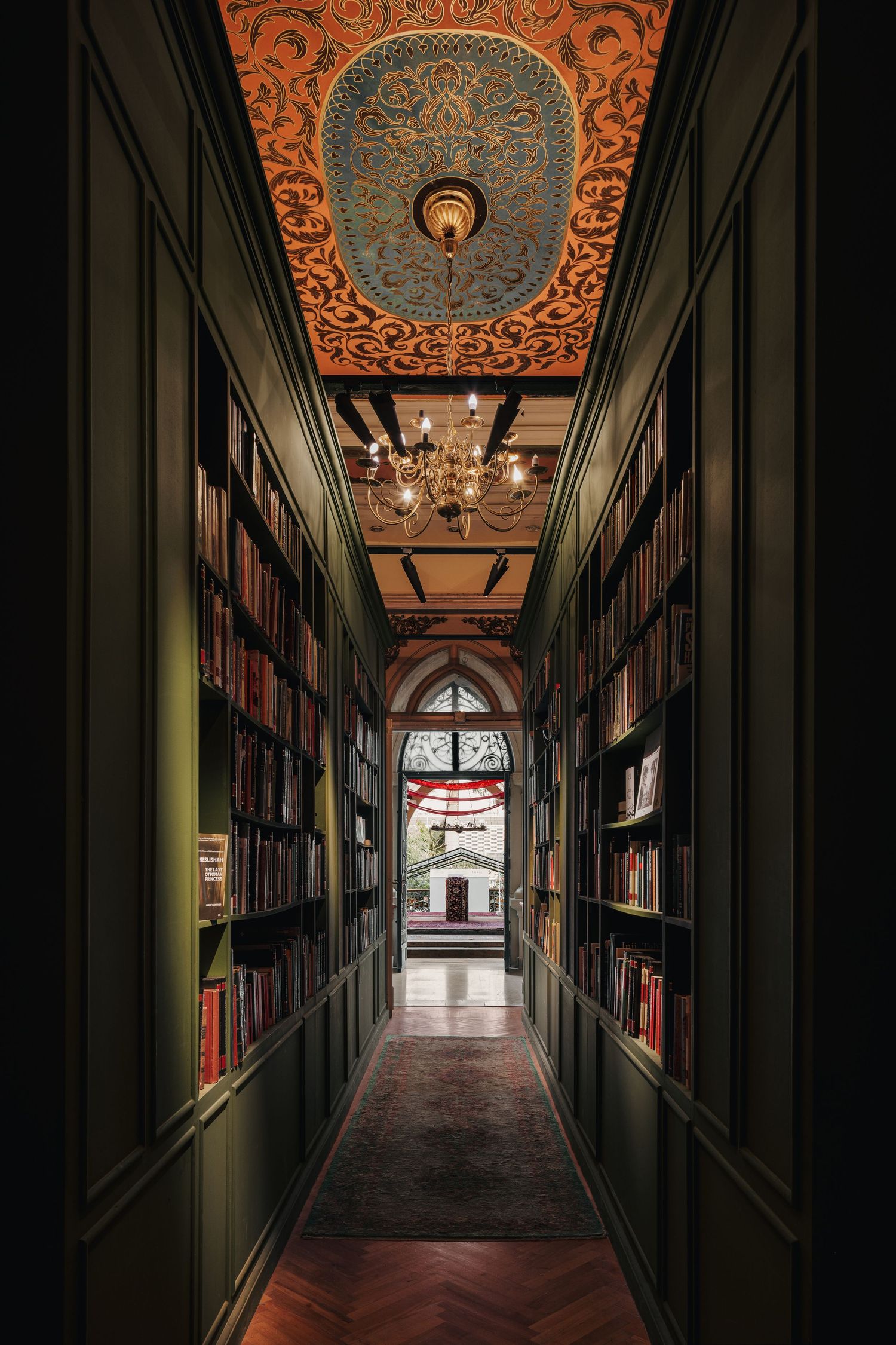 Grand library hallway with floor-to-ceiling bookshelves, ornate painted ceiling, chandelier, and arched window.