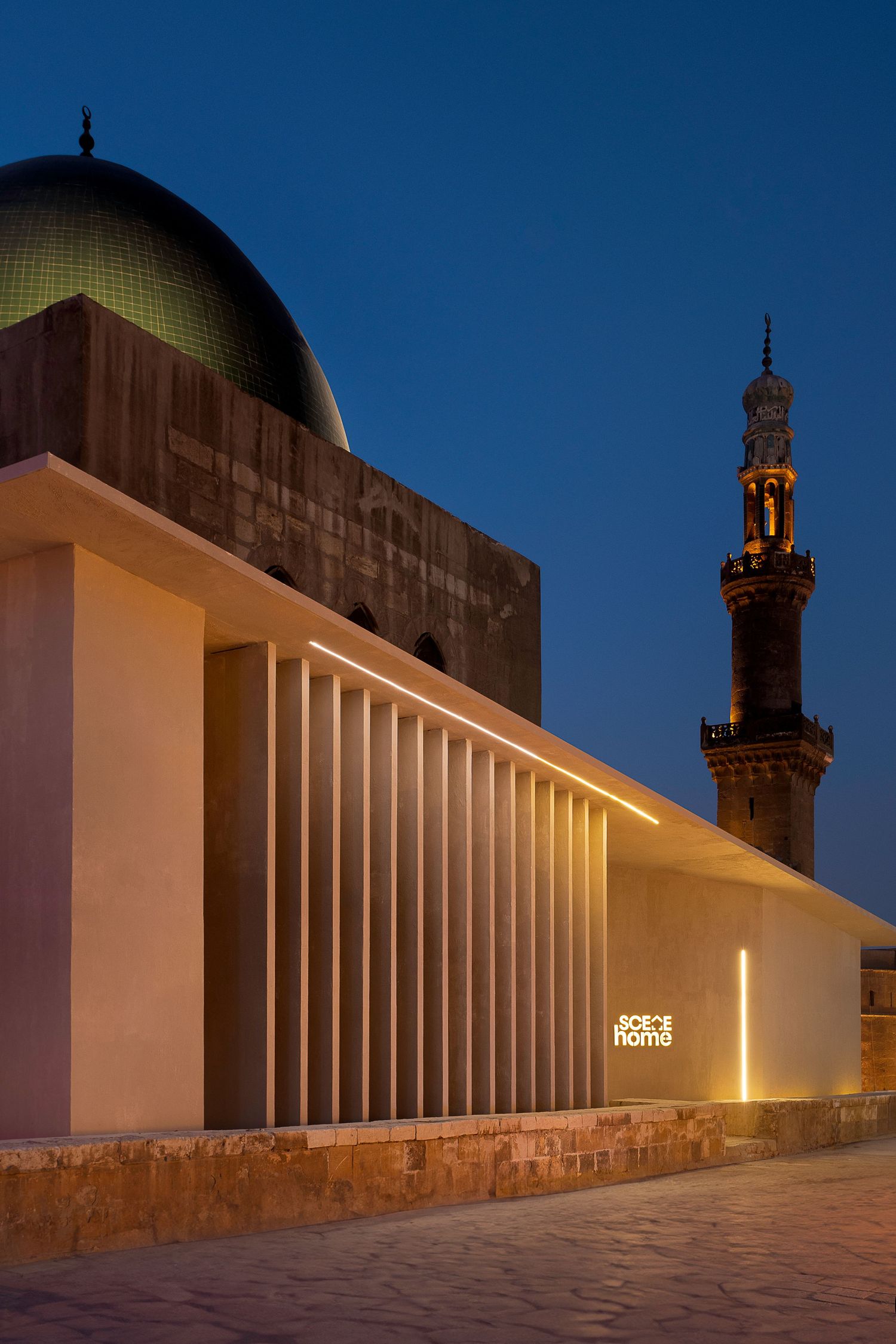 Illuminated modern building facade with vertical fins beside a historic mosque dome and minaret at dusk.