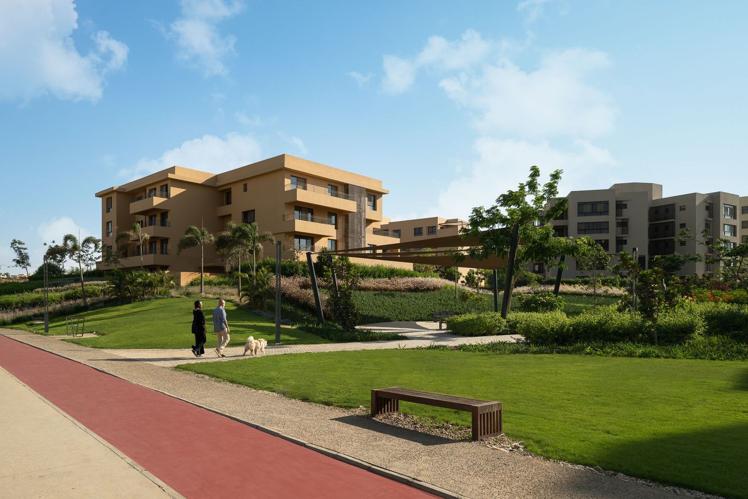 Modern residential complex with lush green lawns, walking paths, and a person walking a dog on a sunny day.