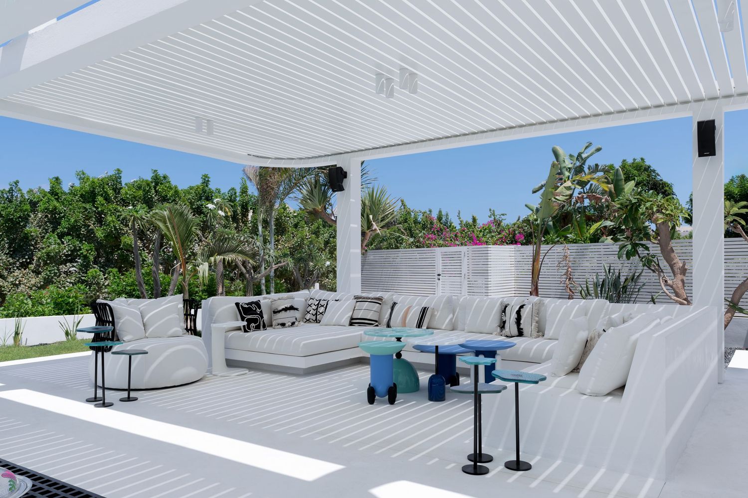 Bright white covered outdoor lounge with sectional sofa, blue accent stools, and lush tropical garden backdrop.