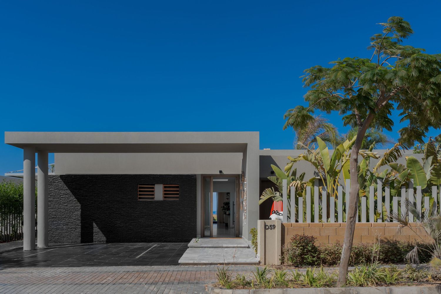 Modern villa entrance with dark brick facade, concrete canopy, white fence, and tropical landscaping.