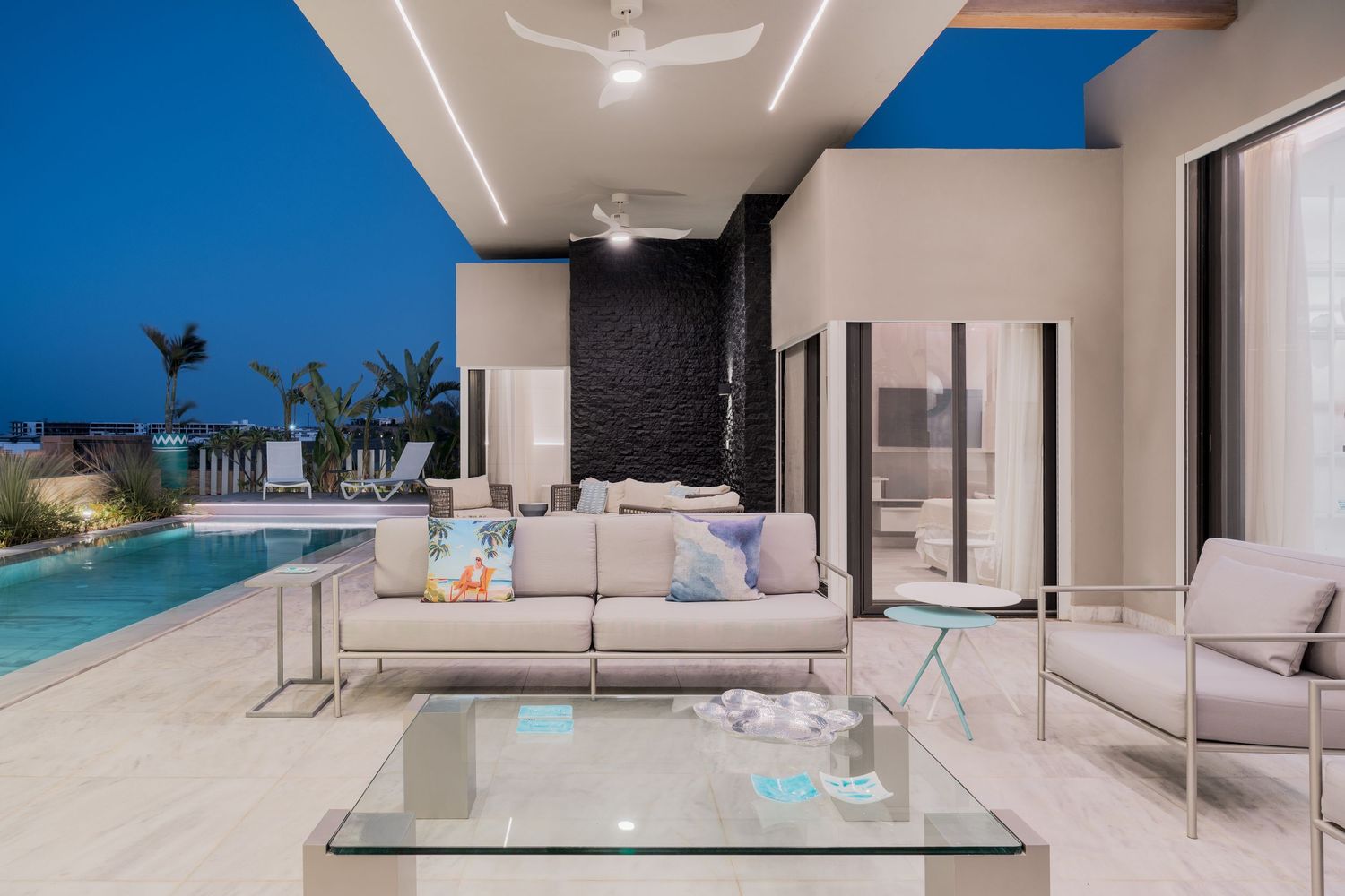 Luxurious poolside living area at dusk with gray sofas, glass coffee table, and illuminated pool.