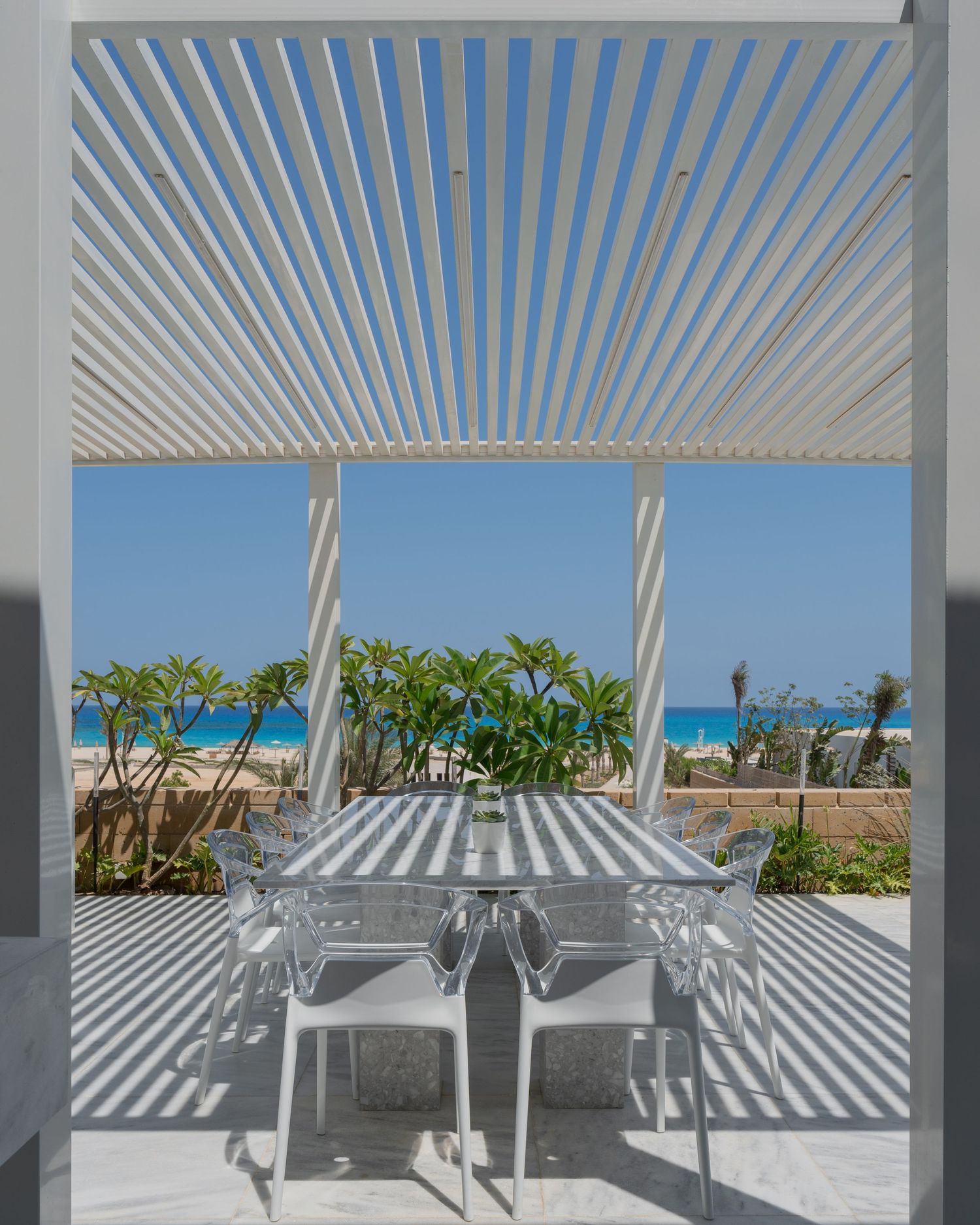 White pergola casting dramatic shadows over outdoor dining table with transparent chairs and sea view.