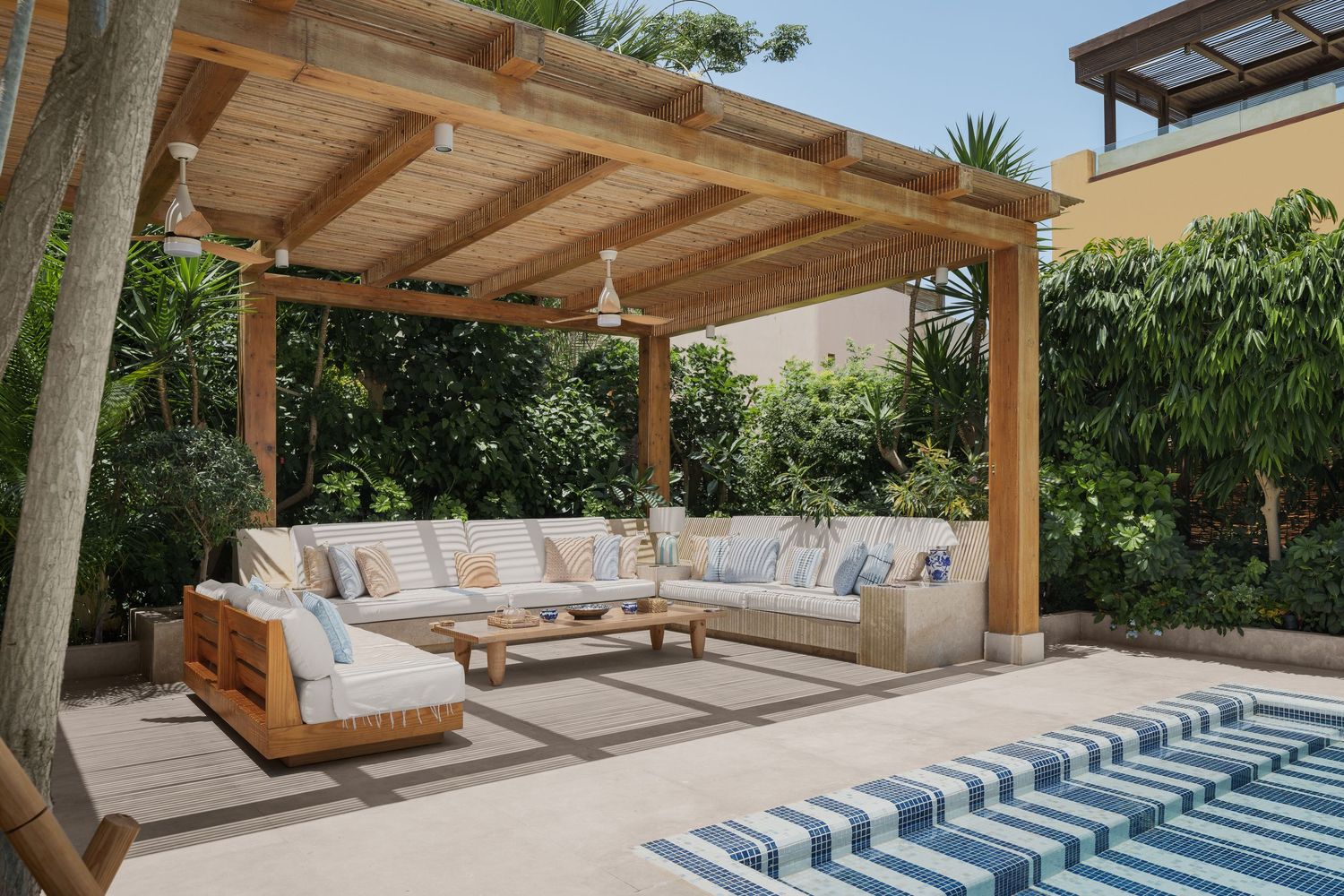 Elegant outdoor lounge area featuring a teak pergola, L-shaped white sofa, and lush garden backdrop.