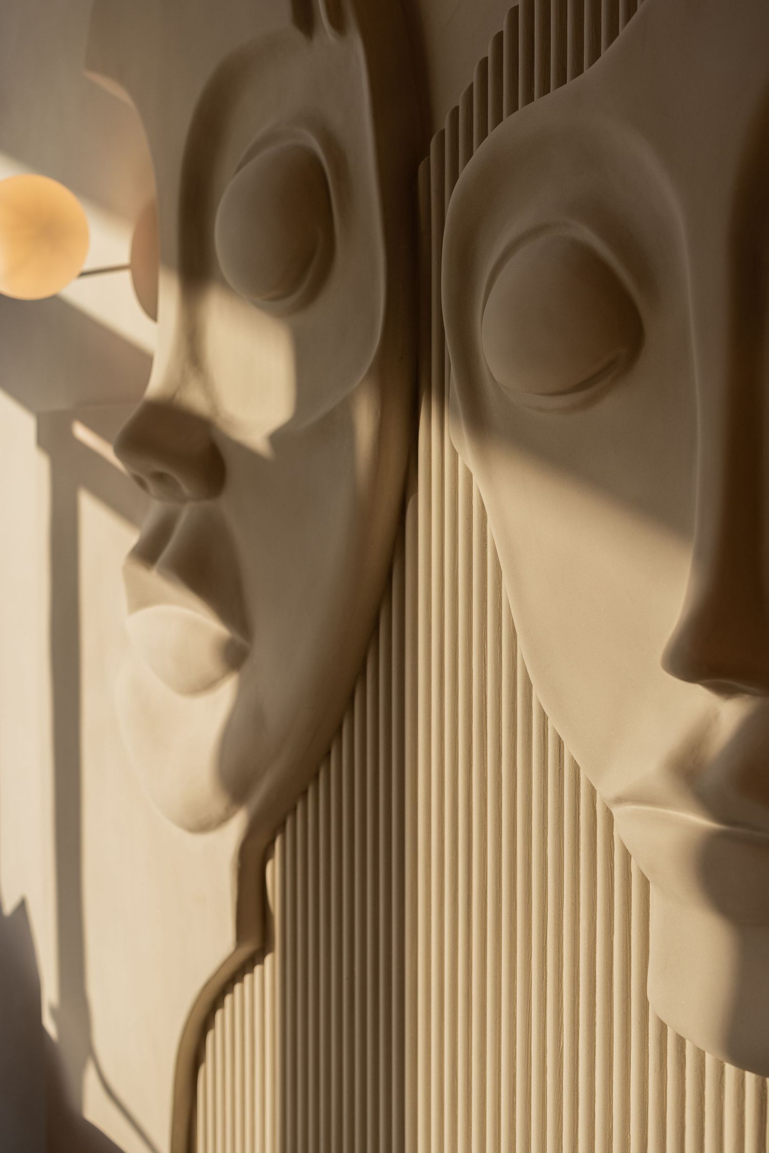 Close-up of two beige sculptural face reliefs on a ribbed wall panel with warm sunlight and shadows.