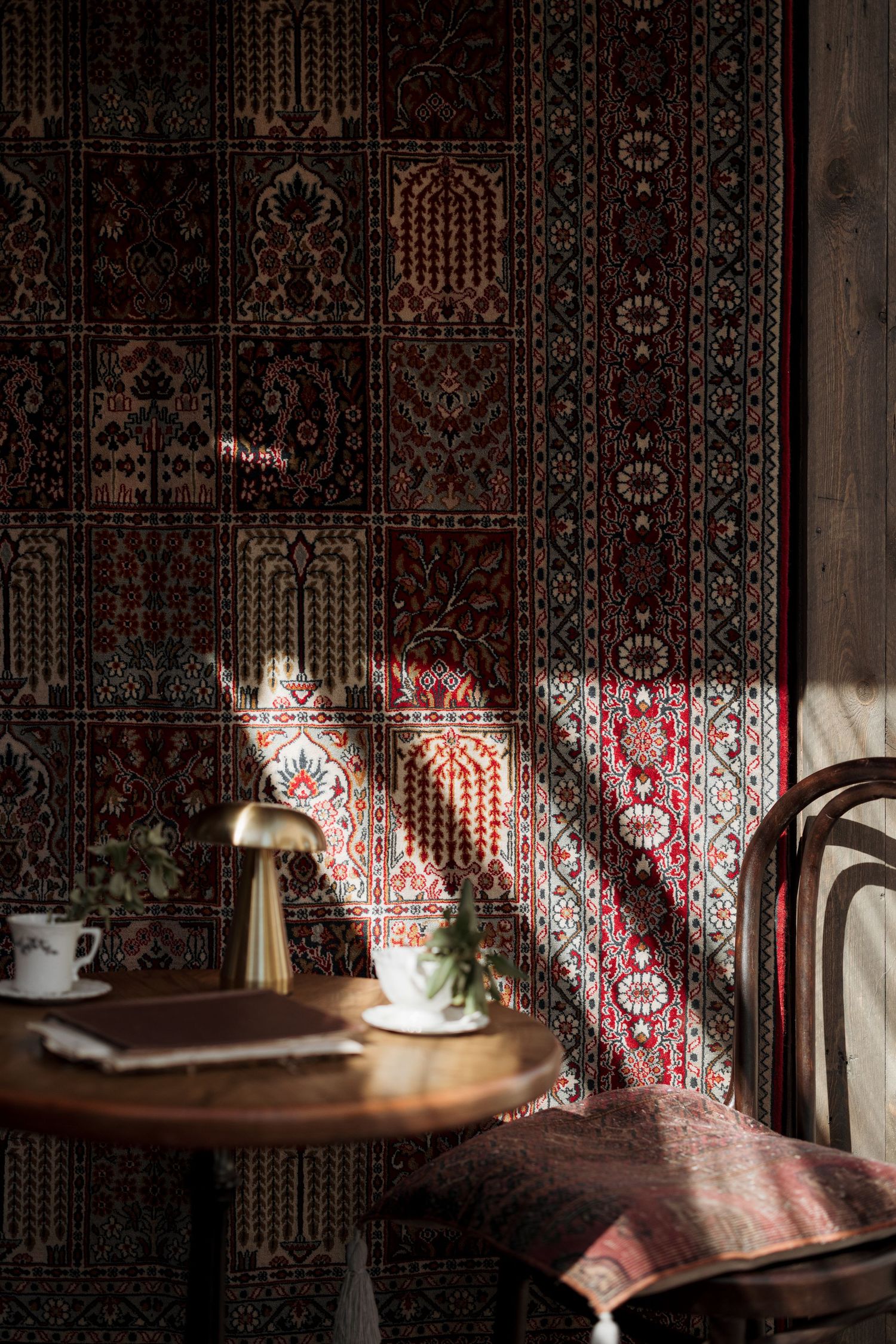 Sunlit cafe corner with ornate dark red Persian carpet wall, wooden table, brass lamp, and vintage chair.