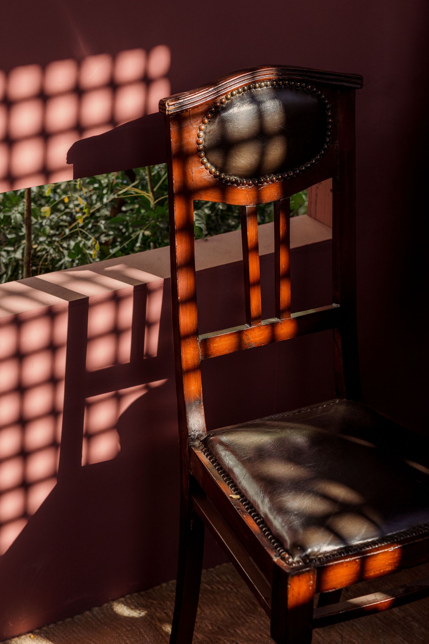 Antique wooden chair with leather seat casting geometric shadow patterns on a dark red wall.
