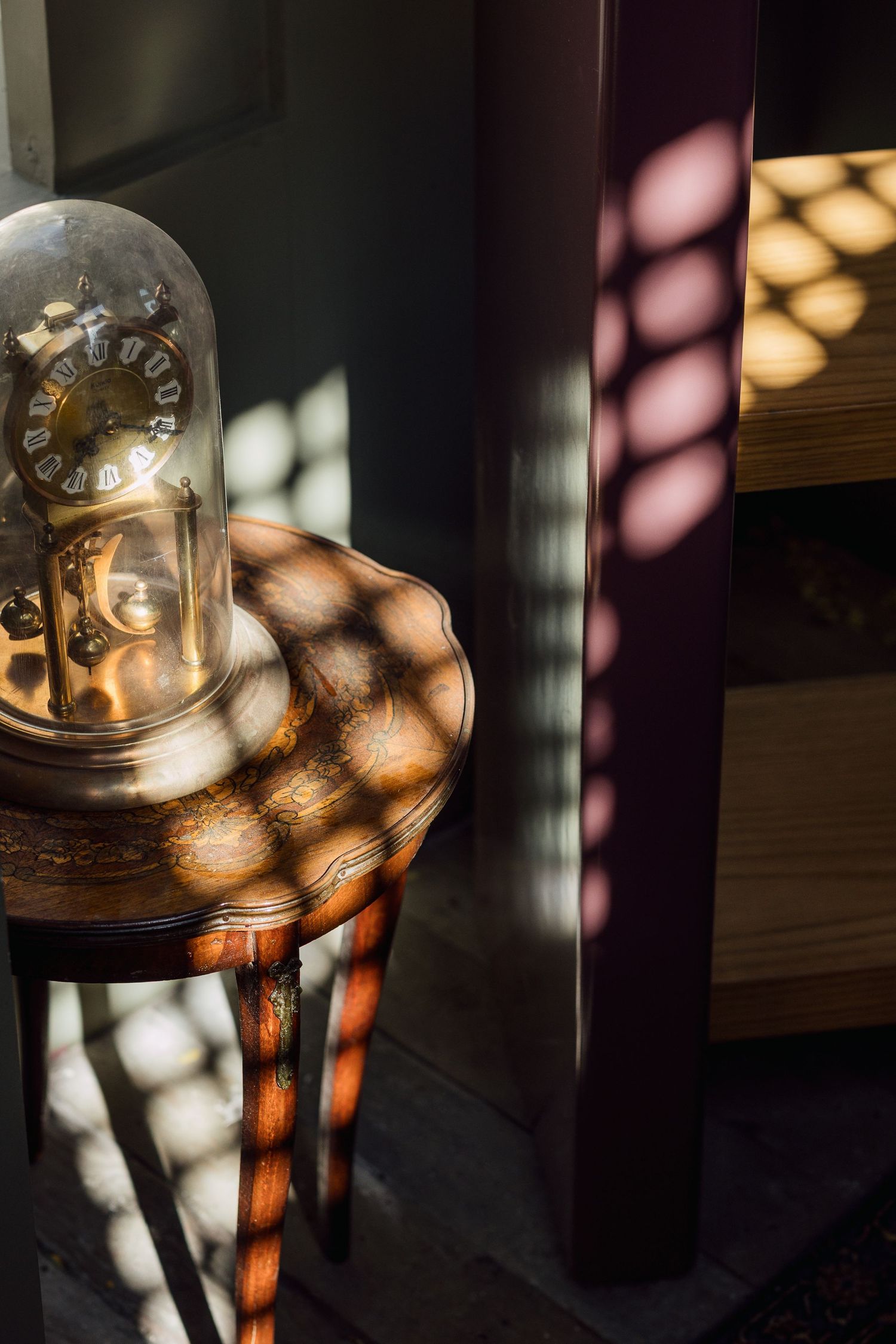 Antique glass dome clock on carved wooden side table with dappled light and shadow patterns.