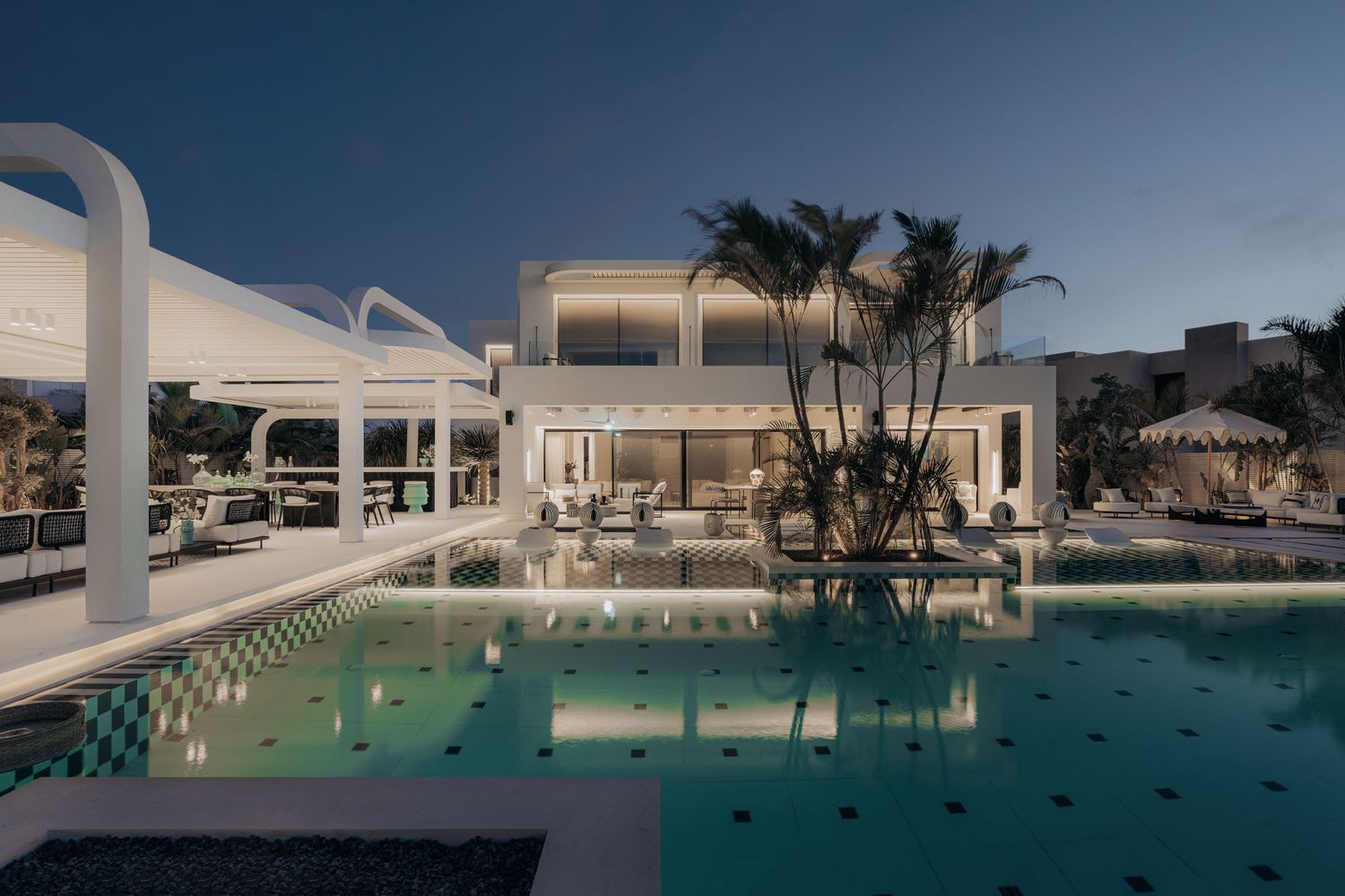 Luxurious white modern villa with illuminated pool at dusk, featuring palm trees and elegant outdoor seating area.