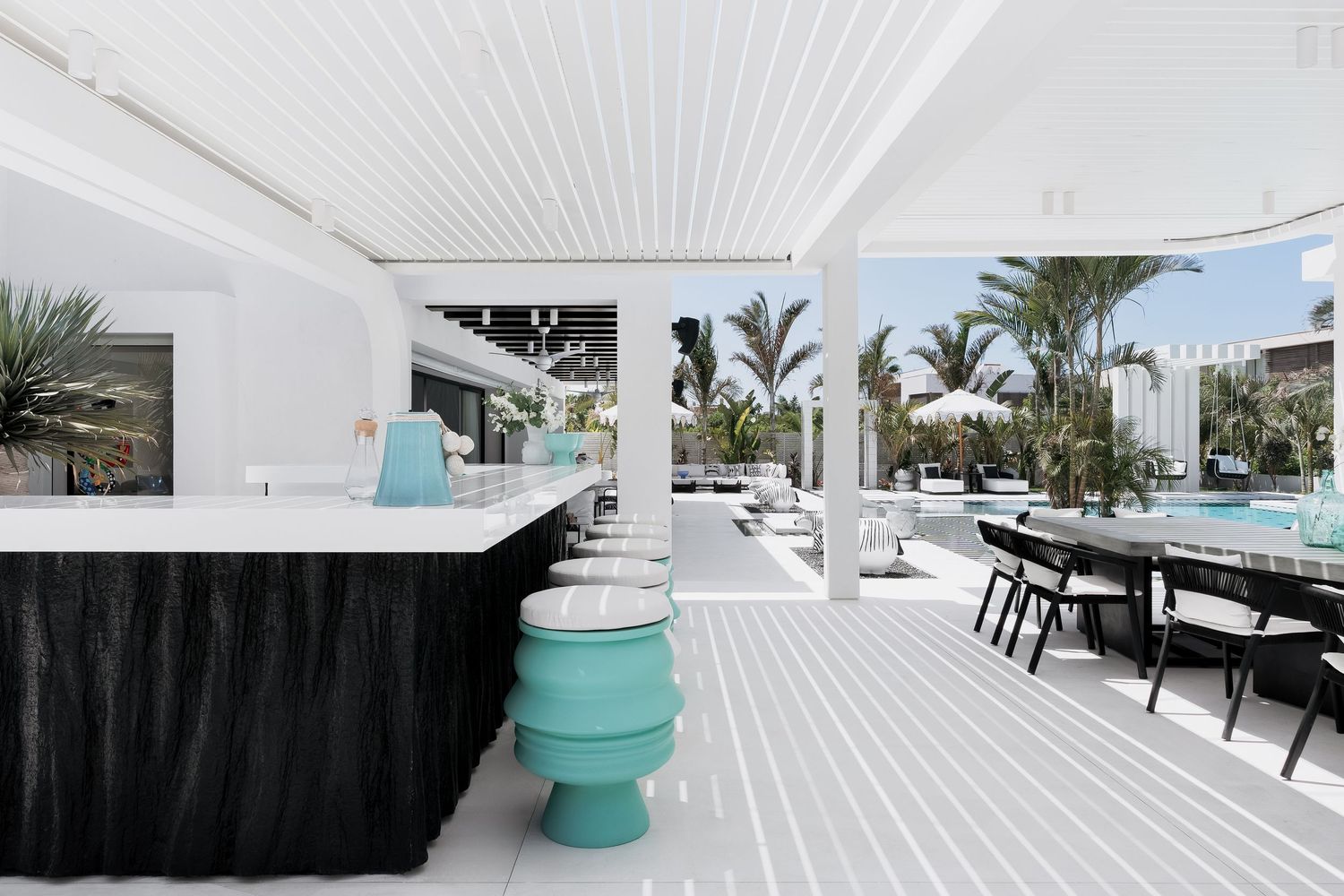 Modern outdoor bar area with black skirted counter, teal stools, white pergola, and pool view surrounded by palm trees.
