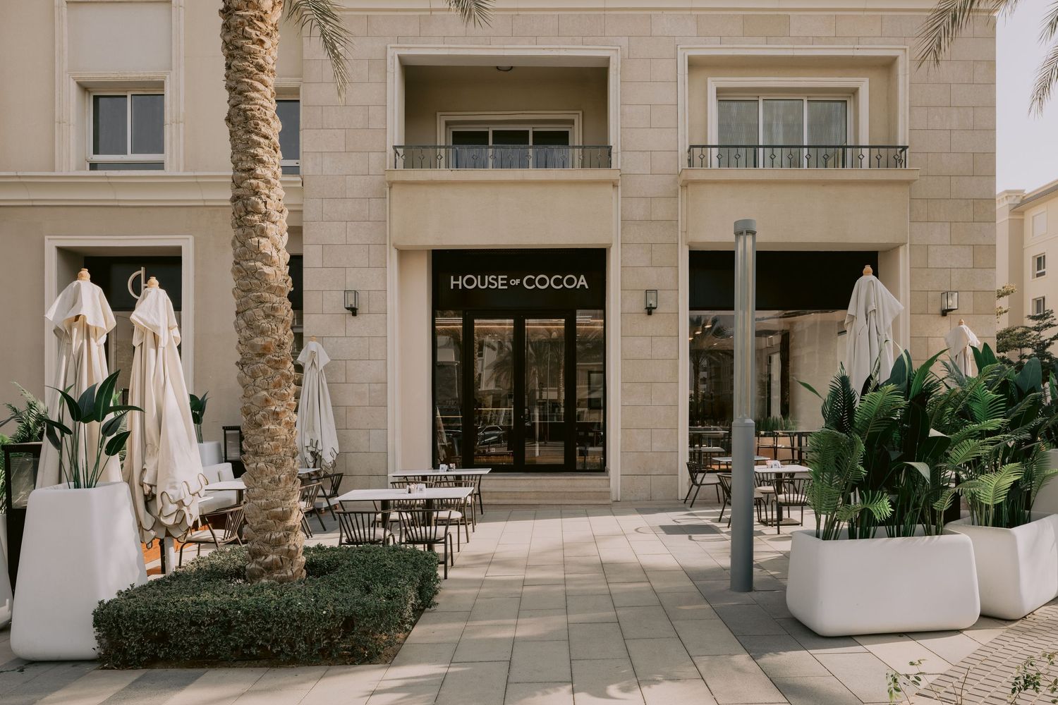 House of Cocoa cafe exterior with outdoor seating, palm trees, and elegant building facade in daylight.