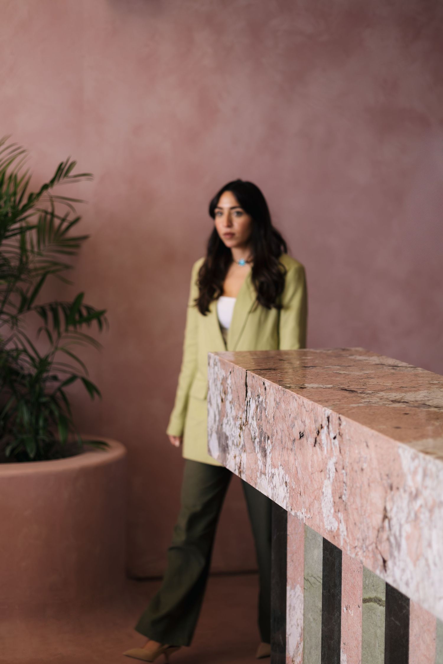 Woman in sage blazer standing beside textured pink travertine marble counter against dusty rose stucco wall.