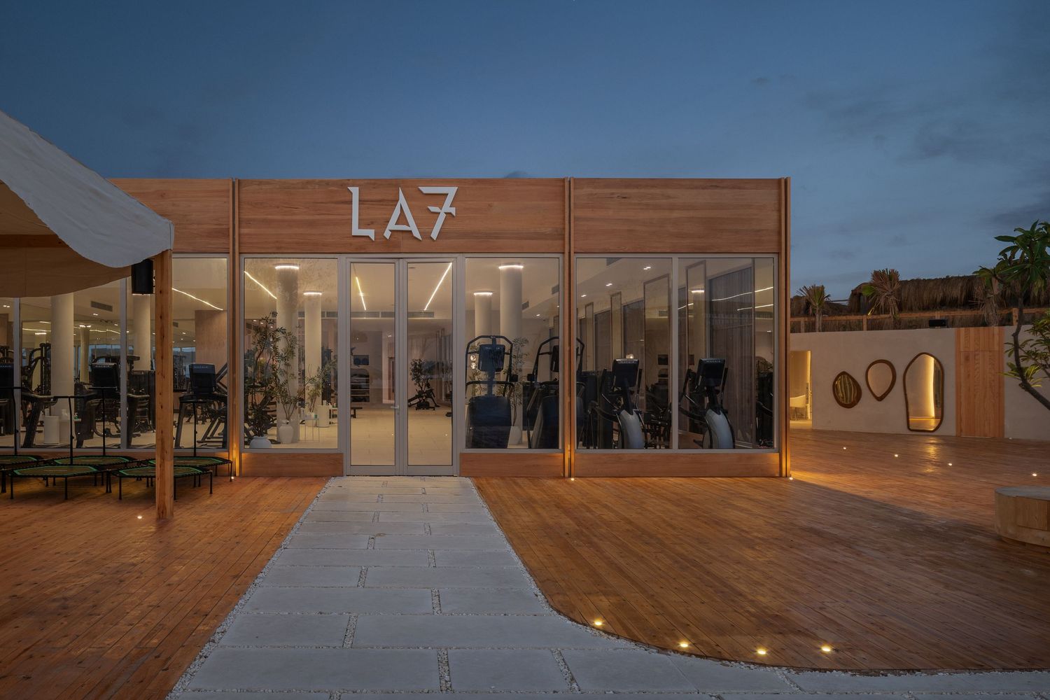 LA7 fitness studio exterior at dusk with warm glowing lights, glass facade, and wooden deck pathway.