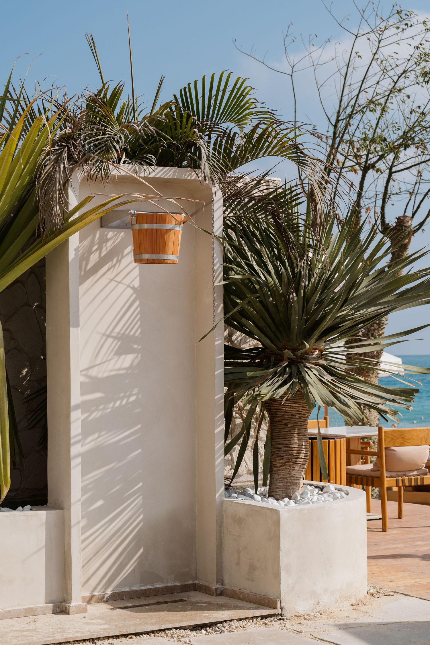 Outdoor beach shower station with wooden bucket, white walls, and tropical palm trees by the sea.