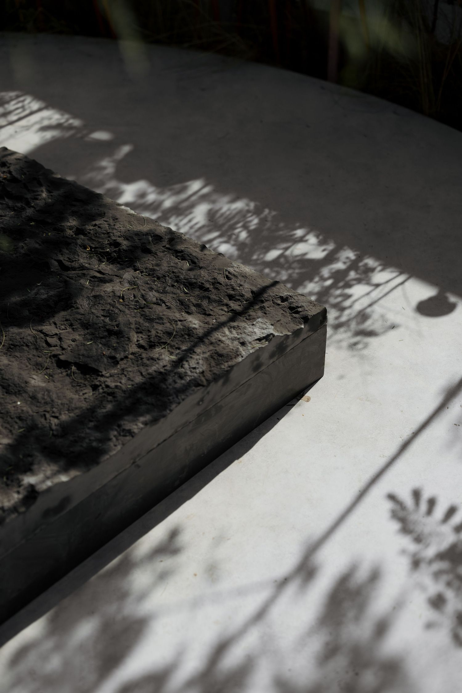 Close-up of a rough dark stone block casting shadows on a white concrete surface outdoors.