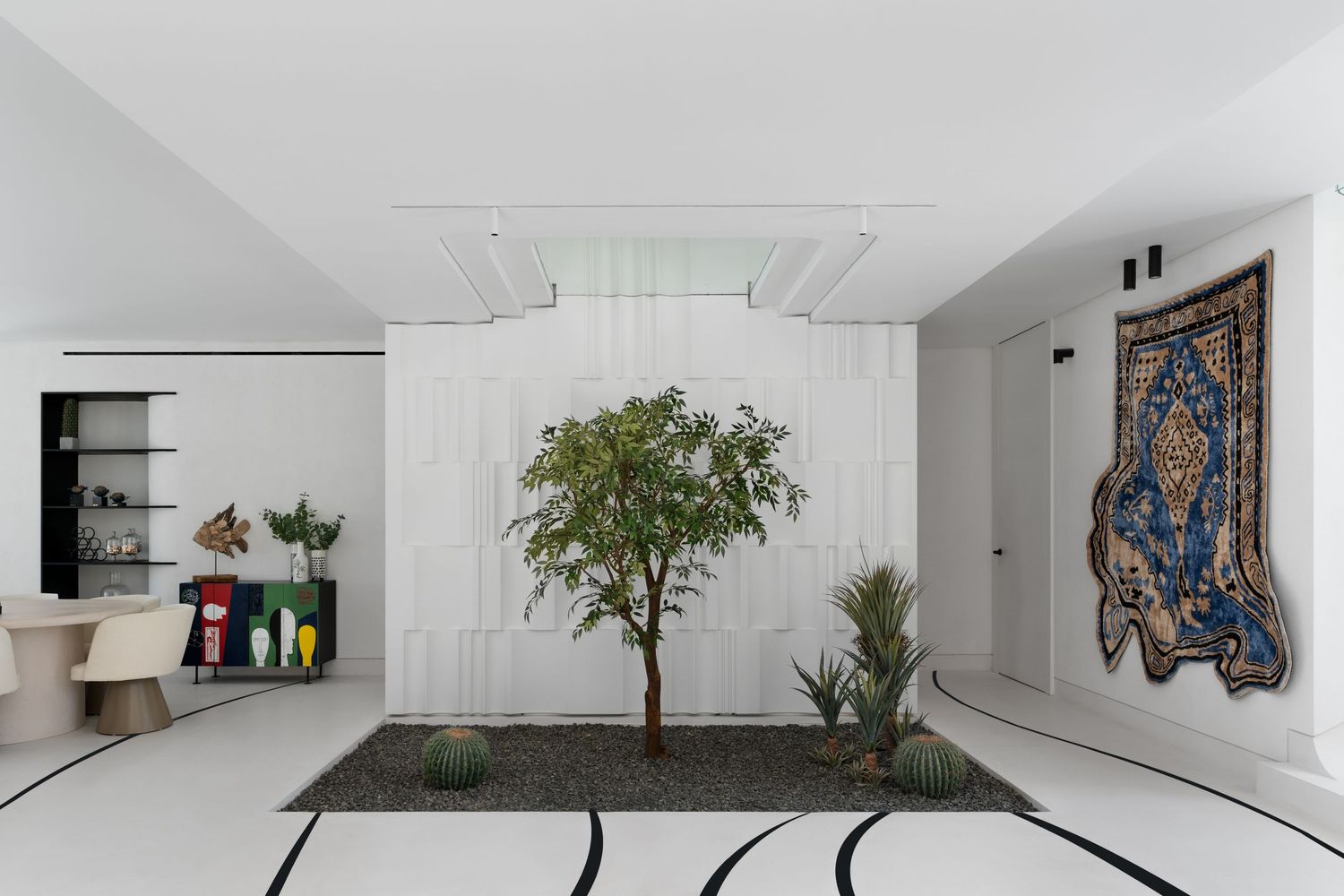 Modern white interior with indoor tree, geometric rug, colorful bookshelf, and ornate blue tapestry wall art.