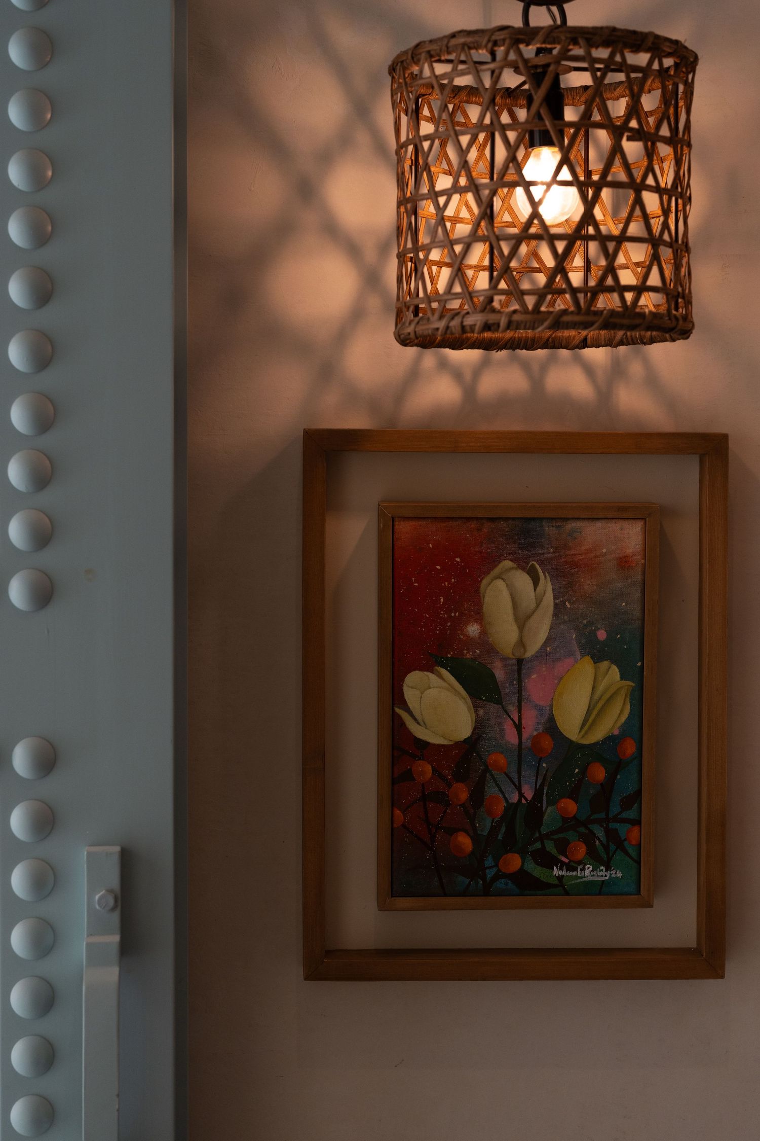 Woven rattan pendant light casting decorative shadows above a framed floral painting on a white wall.