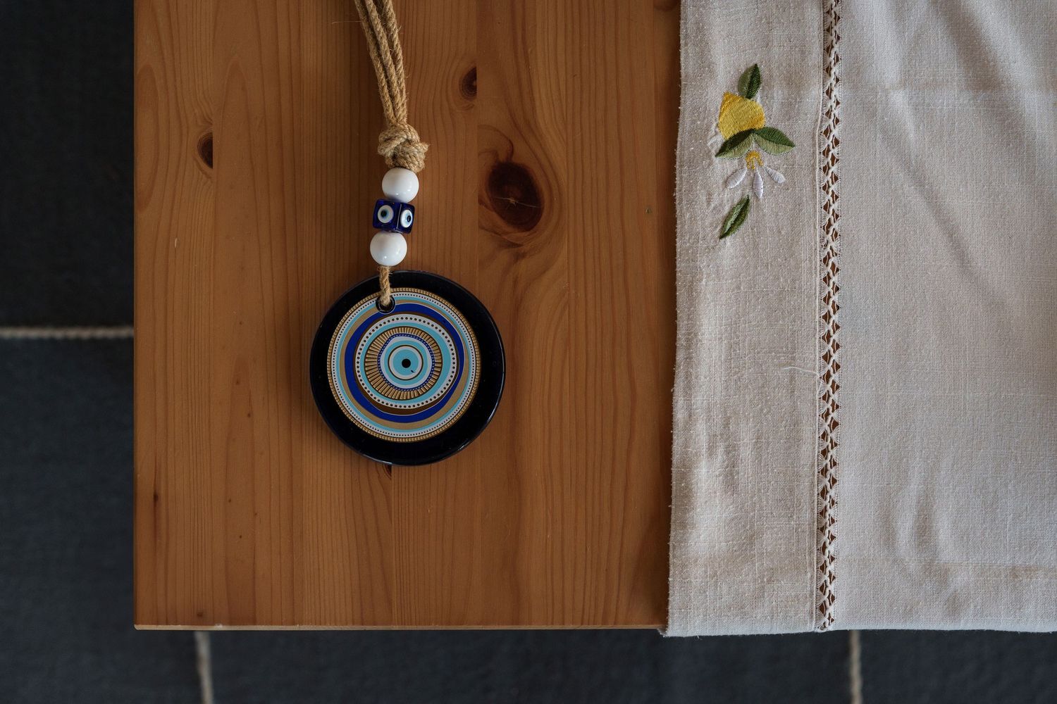Handmade evil eye charm necklace on pine wood surface beside embroidered white linen cloth.