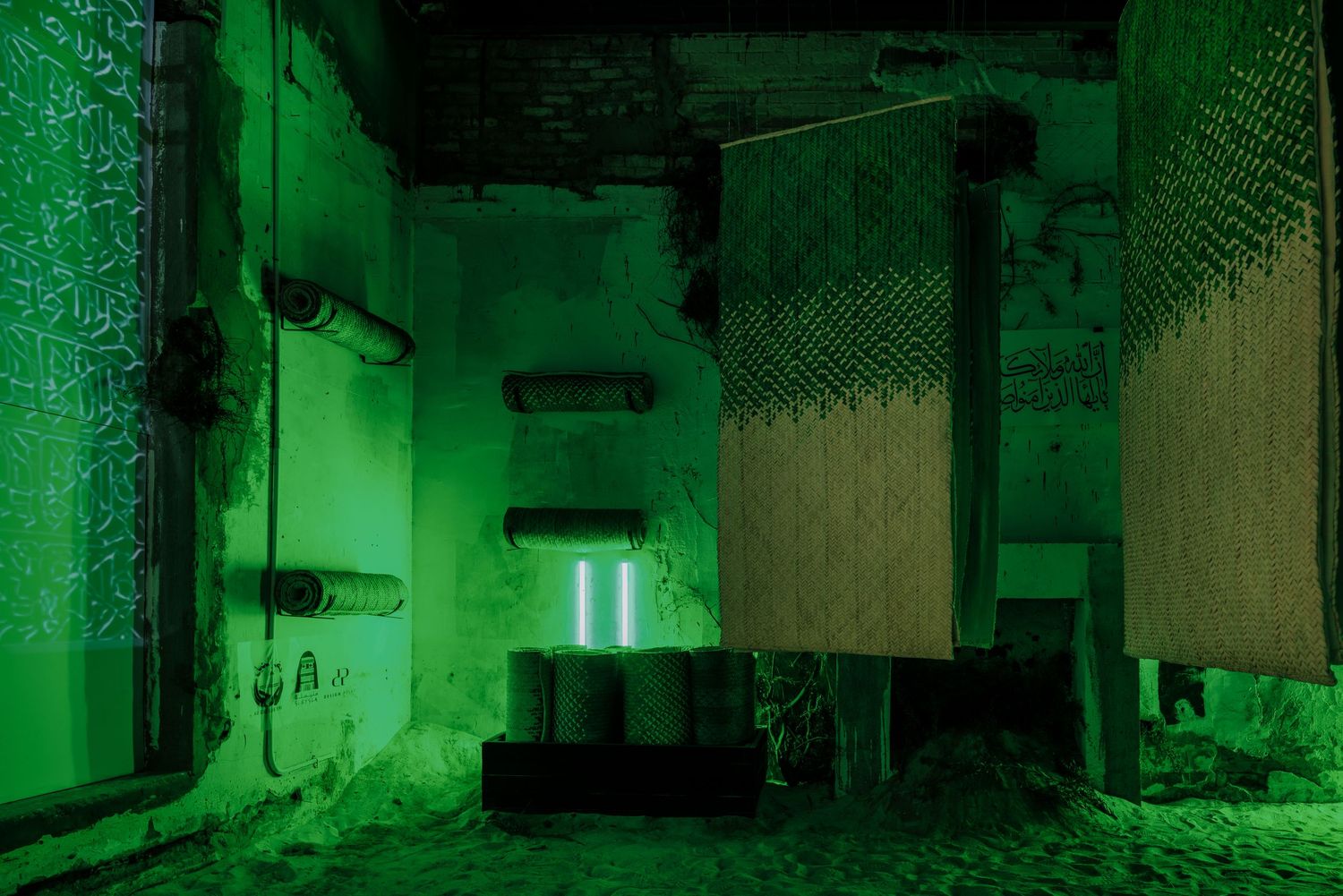 Abandoned industrial space bathed in dramatic green light with hanging textile panels and cylindrical objects.