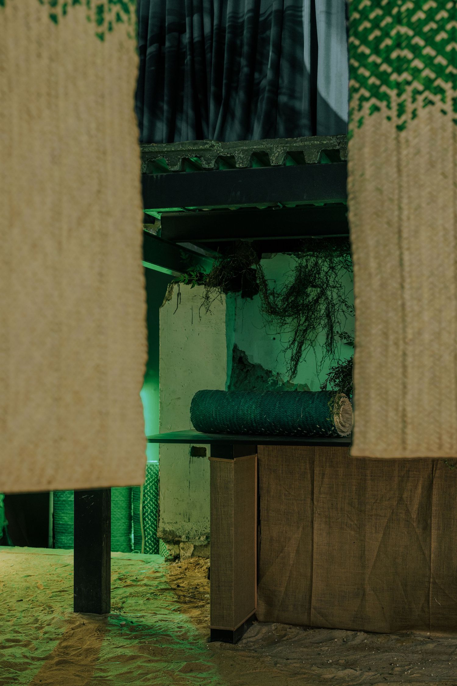 Green-lit abandoned interior viewed through hanging textile panels with sand-covered floor.