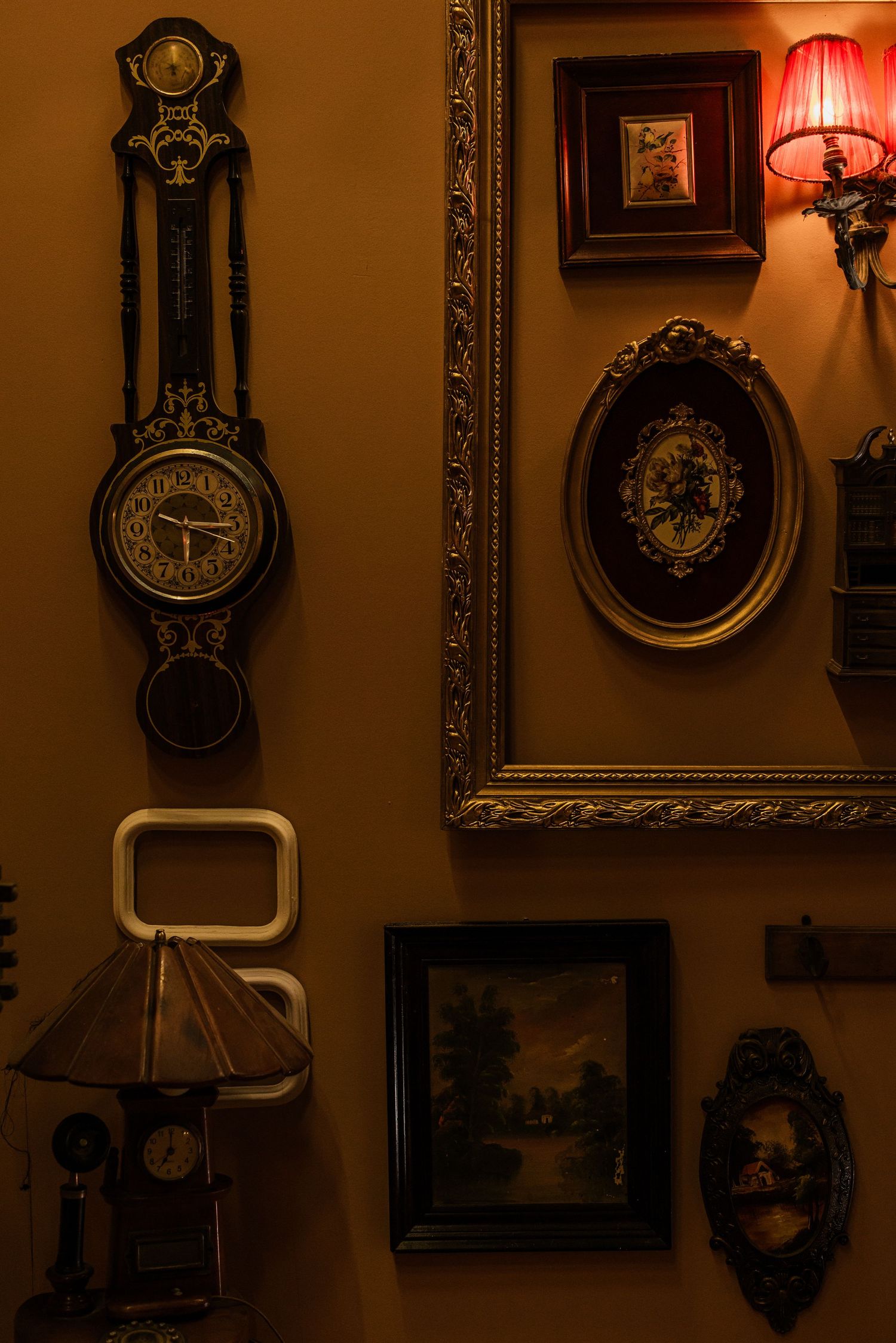 Antique wall clock with ornate frames, vintage paintings, and warm lamp on golden-toned decorative wall.