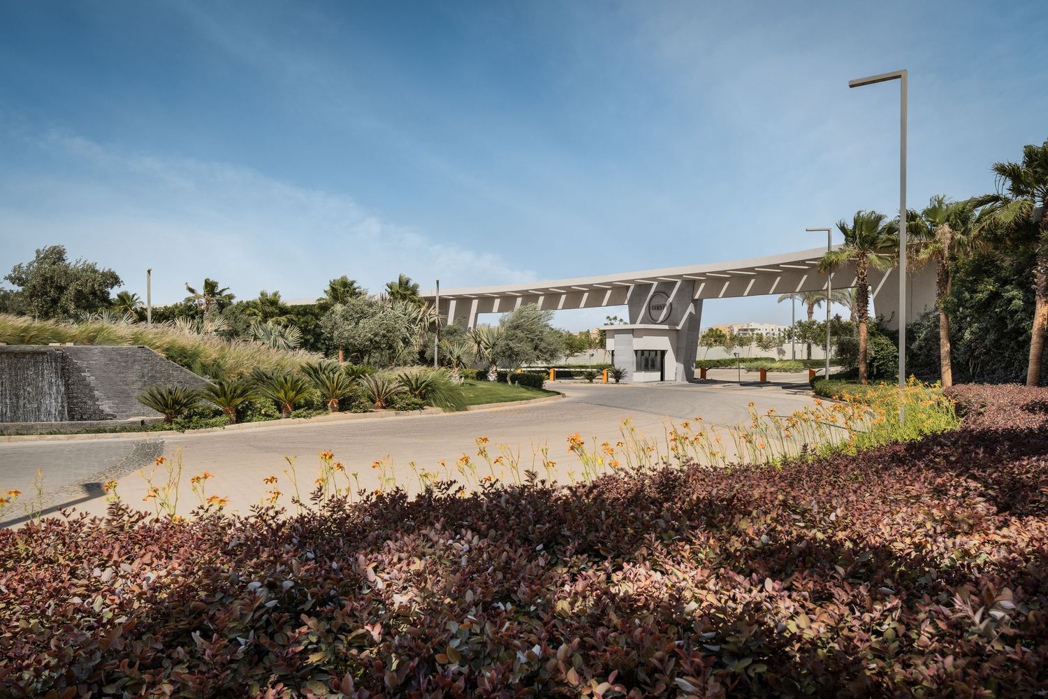 Gated community entrance with modern canopy structure, security booth, and lush red and green landscaping.