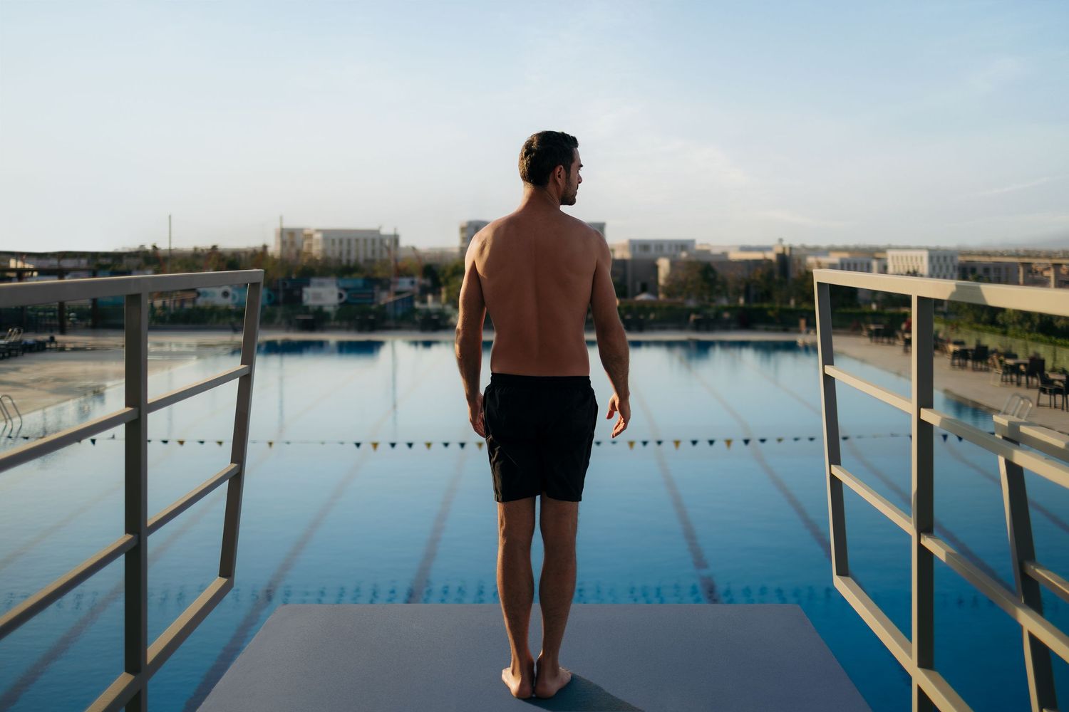 Athletic man standing shirtless on a diving platform overlooking a large outdoor swimming pool at sunset.