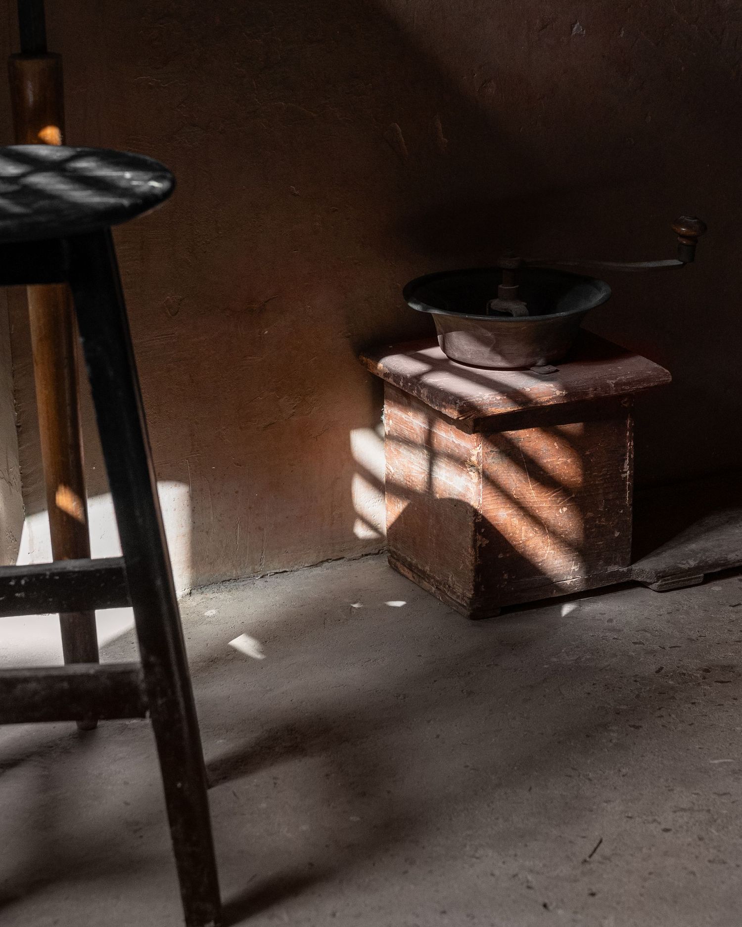 Vintage manual coffee grinder on a wooden box with dramatic sunlight casting shadows on a bare wall.